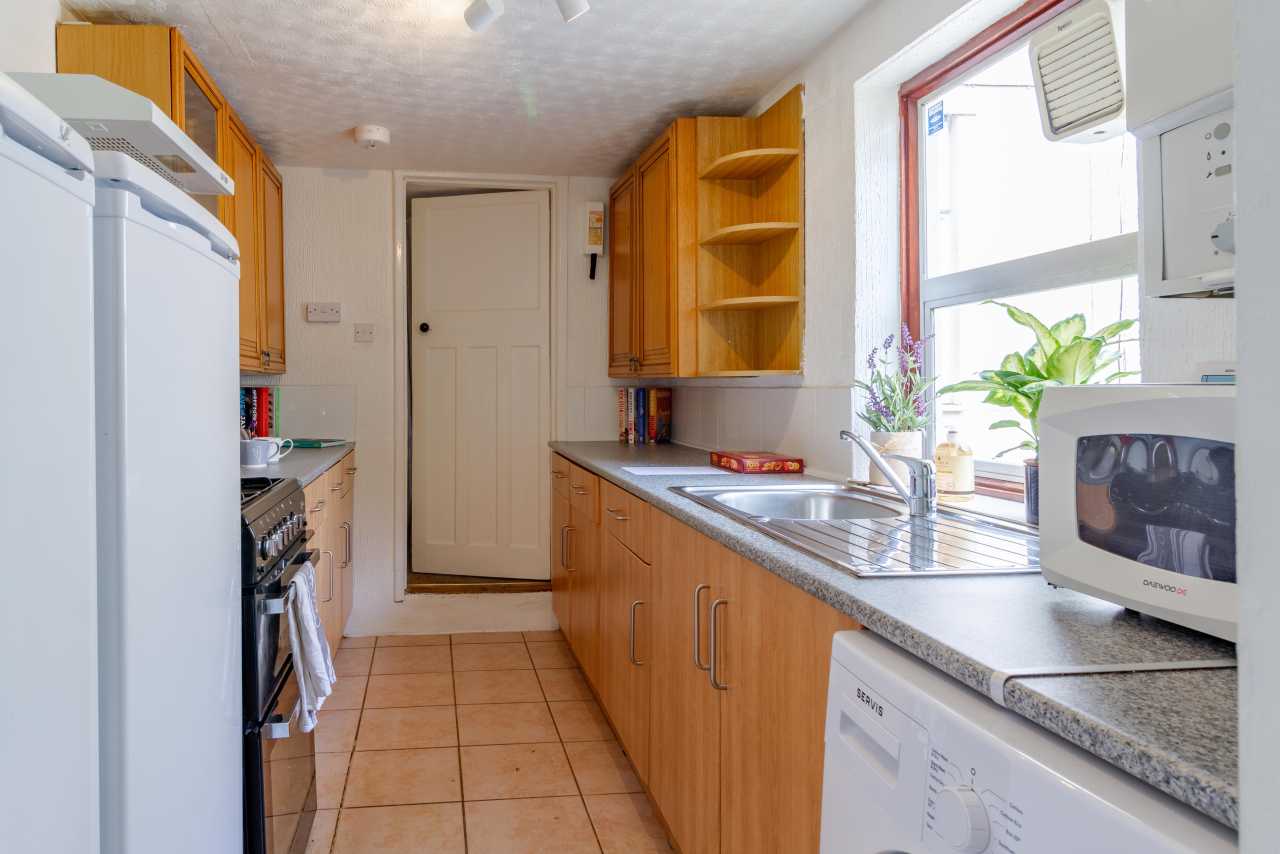 A narrow kitchen with wooden cabinets, tiled floor, cooker, sink, dishwasher, refrigerator and a window.