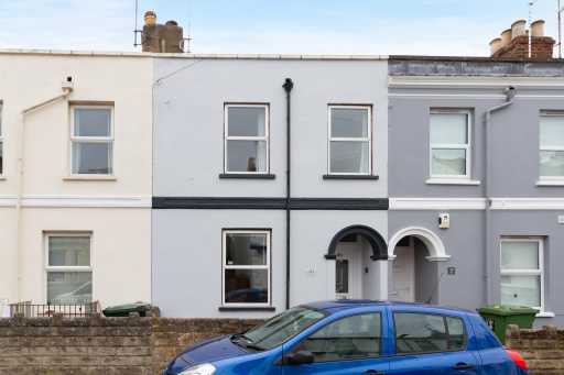 Street view of adjoining terraced houses with painted façades, windows, arched entrances and a blue car in front.