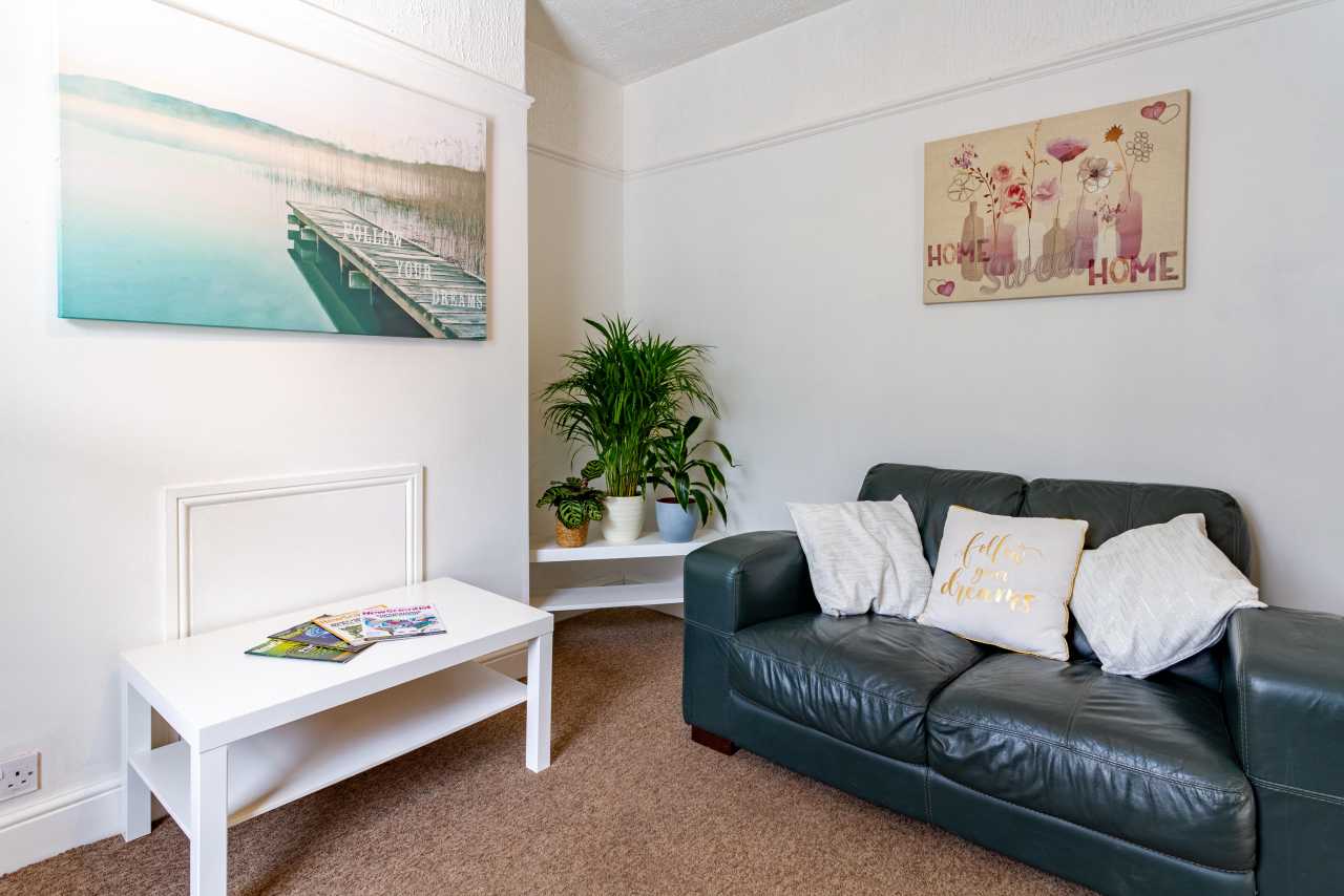 Bright sitting room with a dark sofa, white table, potted plants, wall art and carpeted floor.