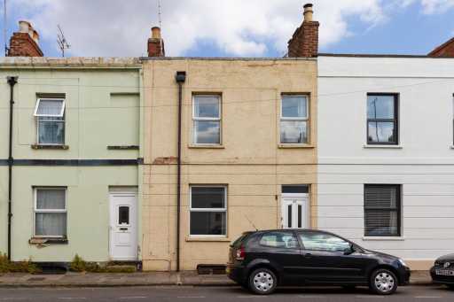 A row of attached terraced houses with brick façades, windows, doors, chimneys, and parked cars along the street.