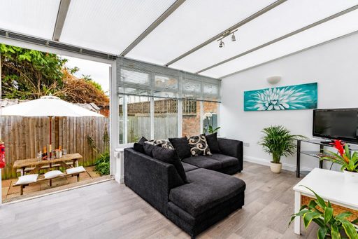 Bright enclosed living area with a dark sectional sofa, television, plants and wall art, opening onto a fenced patio with outdoor seating.