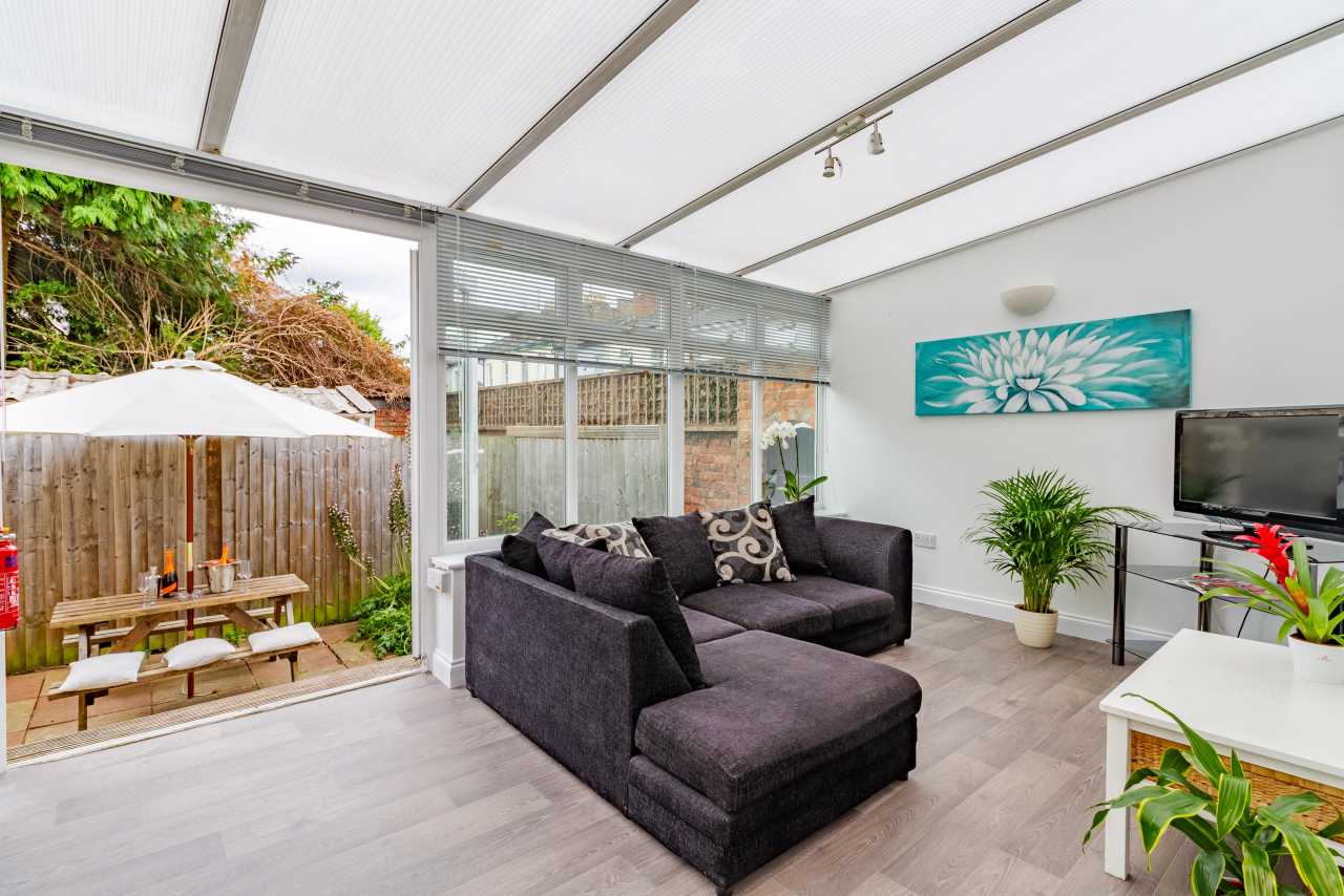 Bright enclosed living area with a dark sectional sofa, television, plants and wall art, opening onto a fenced patio with outdoor seating.