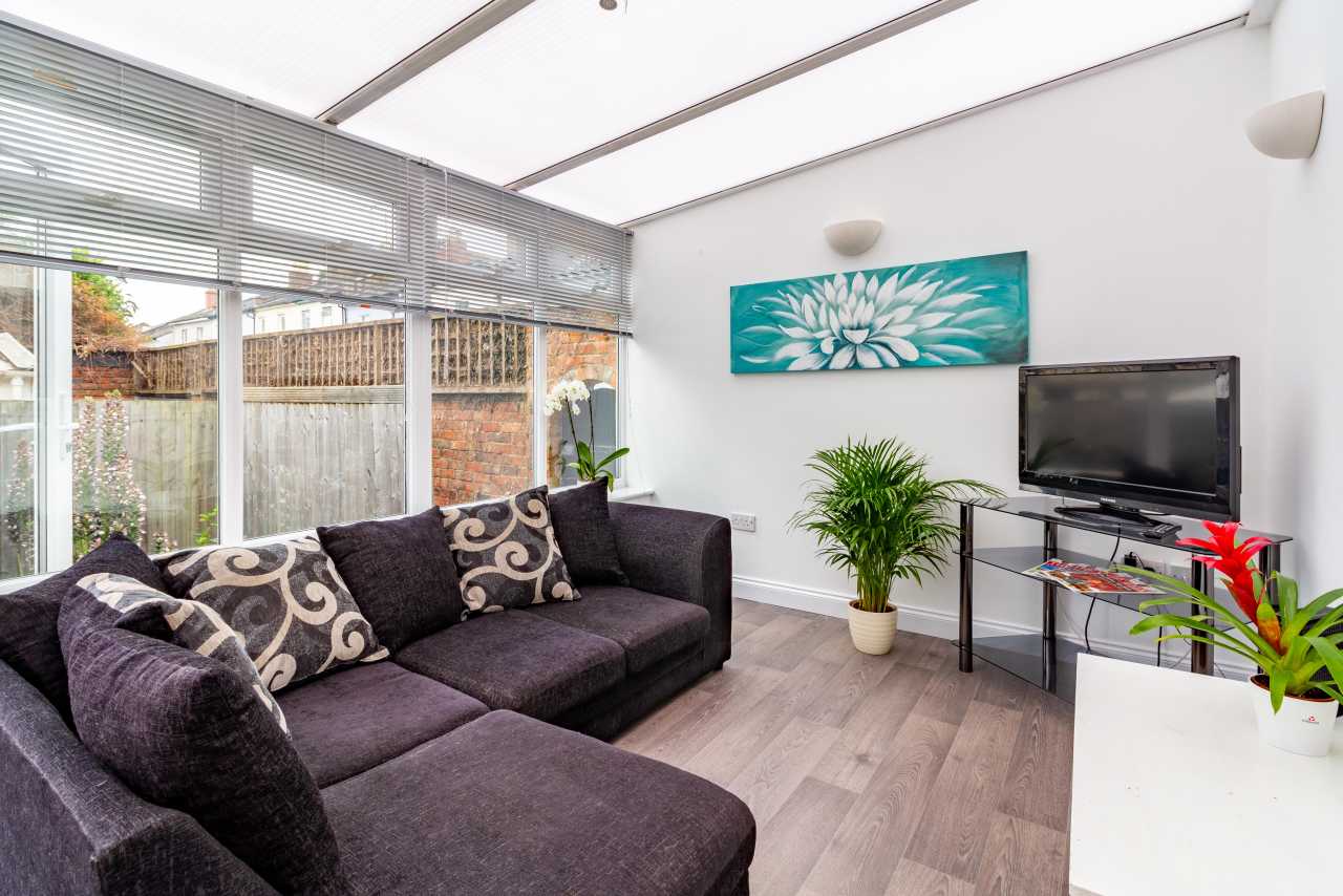Bright furnished living room with a sectional sofa, television, wall art, plants and large windows.