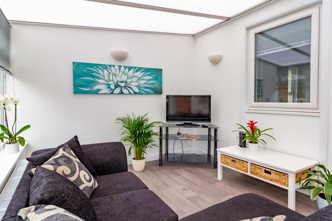 Bright furnished sitting area with a corner sofa, television, plants, wall art and a white storage cabinet.