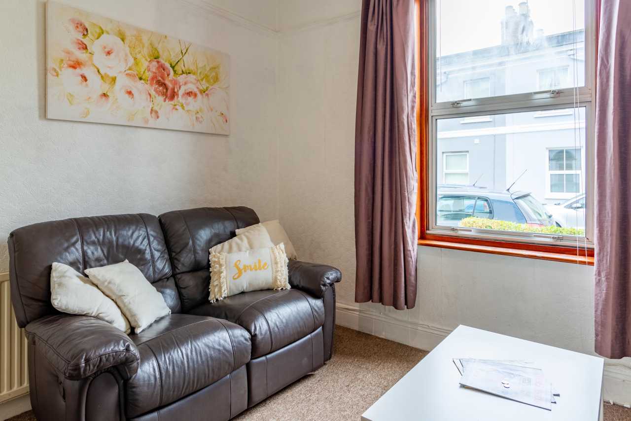 A sitting area with a dark leather sofa, cushions, floral wall art, curtains, a window, and a white coffee table.