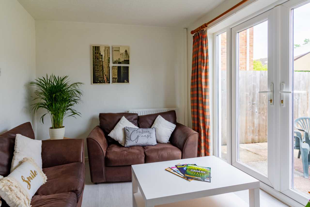 Bright living room with two brown sofas, a white coffee table, a potted plant, wall art and large glass doors opening to a patio.