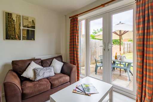 A furnished living area with a brown sofa, white coffee table, framed wall art and glass doors opening onto a patio and garden.