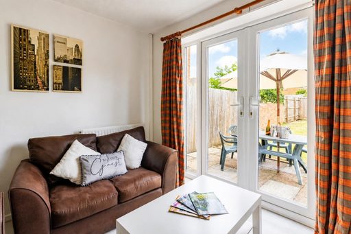 A furnished living area with a brown sofa, white coffee table, framed wall art and glass doors opening onto a patio and garden.