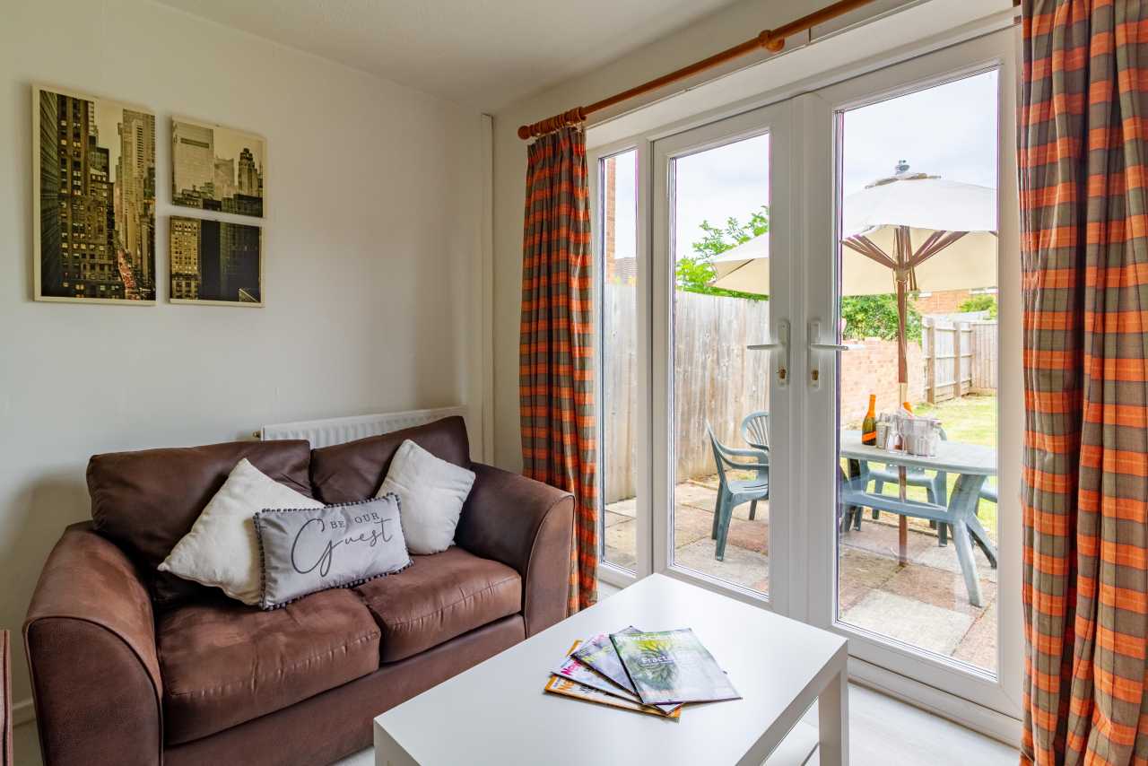 A furnished living area with a brown sofa, white coffee table, framed wall art and glass doors opening onto a patio and garden.