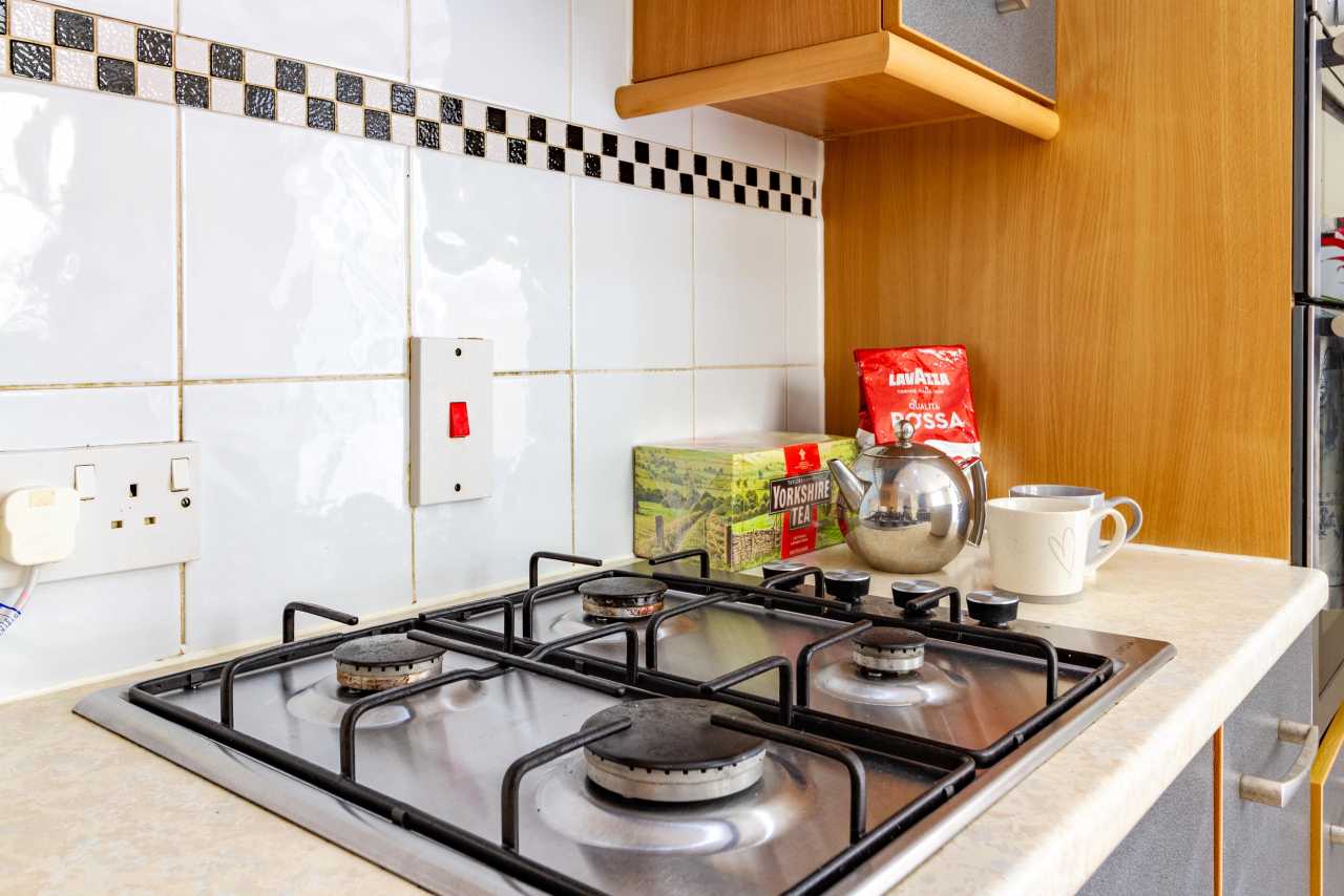 Close-up of a four-burner gas hob on a kitchen countertop, with tiled walls, cupboards, a kettle and a mug.