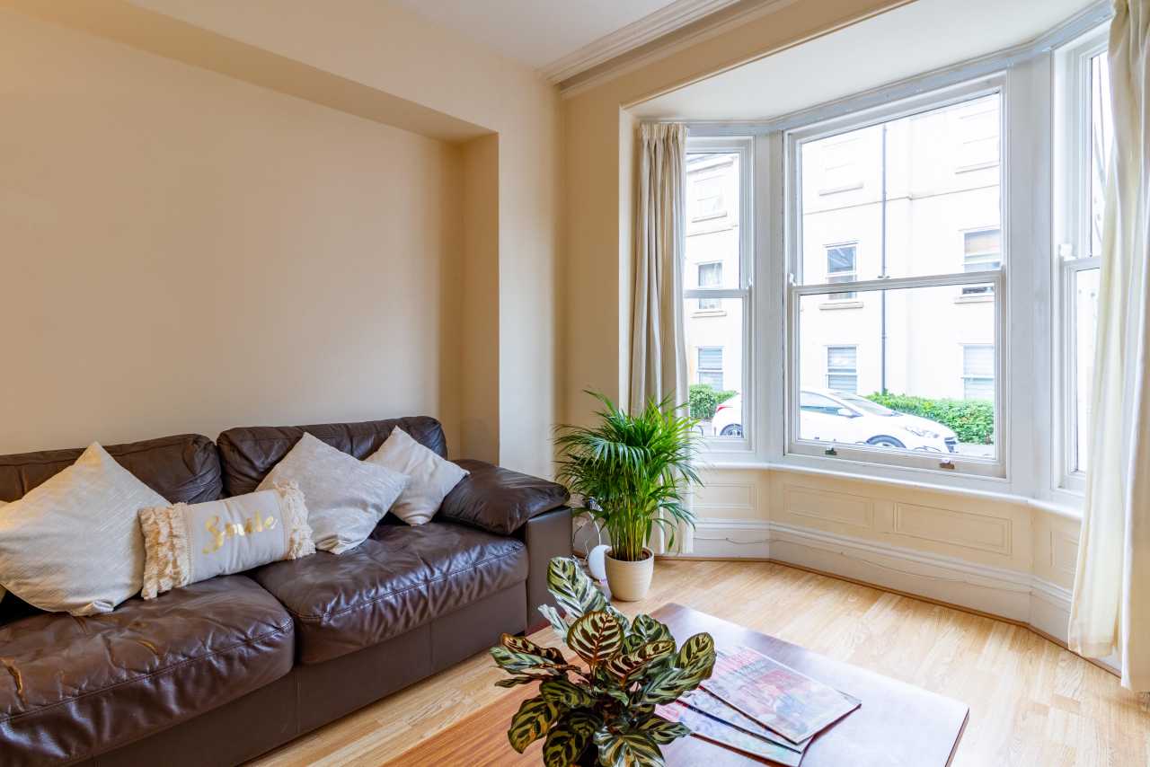Bright living room with a brown leather sofa, cushions, plants, a rug and large bay windows.