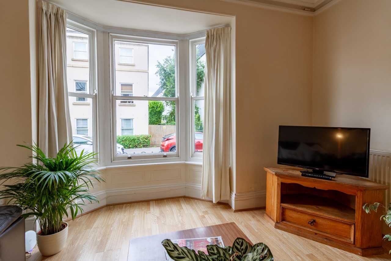 Bright furnished sitting room with a bay window, curtains, television on a wooden stand, wooden flooring and a potted plant.