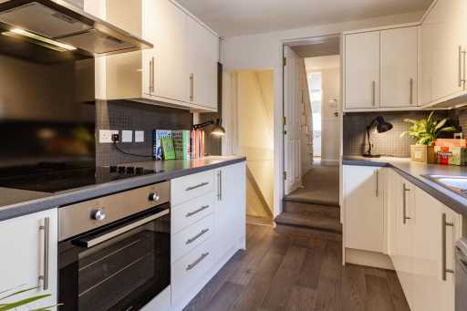 Modern fitted kitchen with white cabinets, built-in oven and hob, tiled backsplash, and a doorway leading to carpeted stairs.