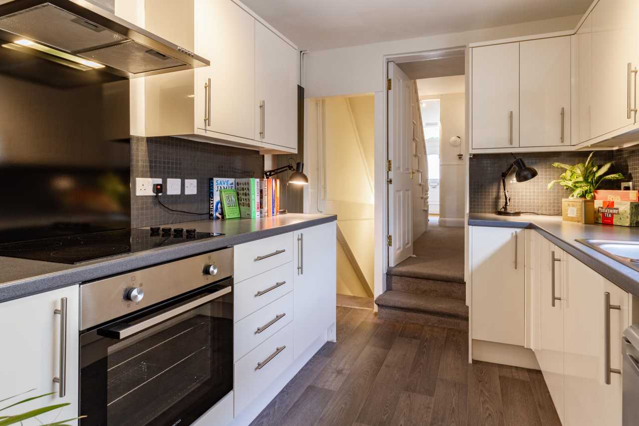 Modern fitted kitchen with white cabinets, built-in oven and hob, tiled backsplash, and a doorway leading to carpeted stairs.