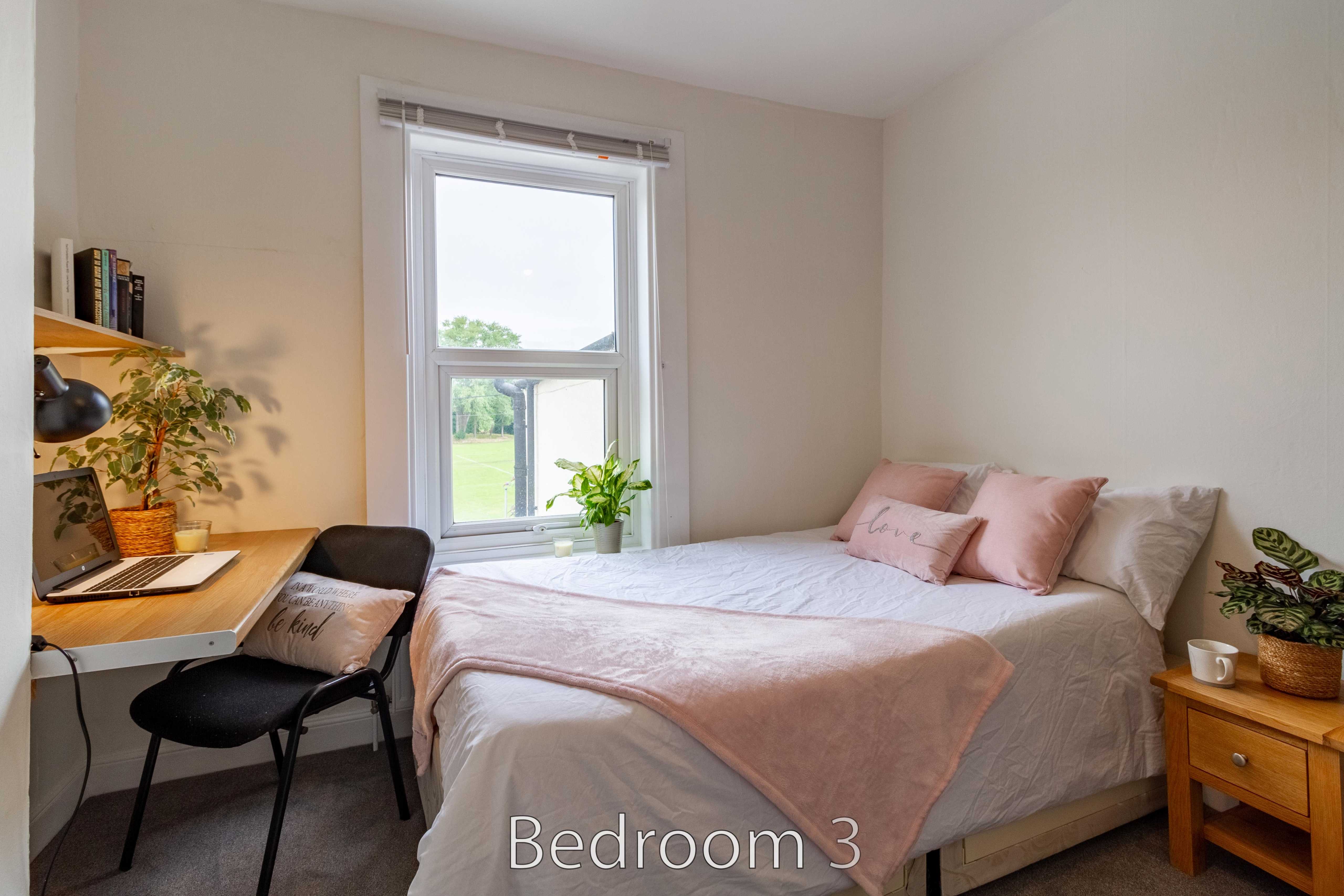 A furnished bedroom with a double bed, desk, chair, window, and bedside table.