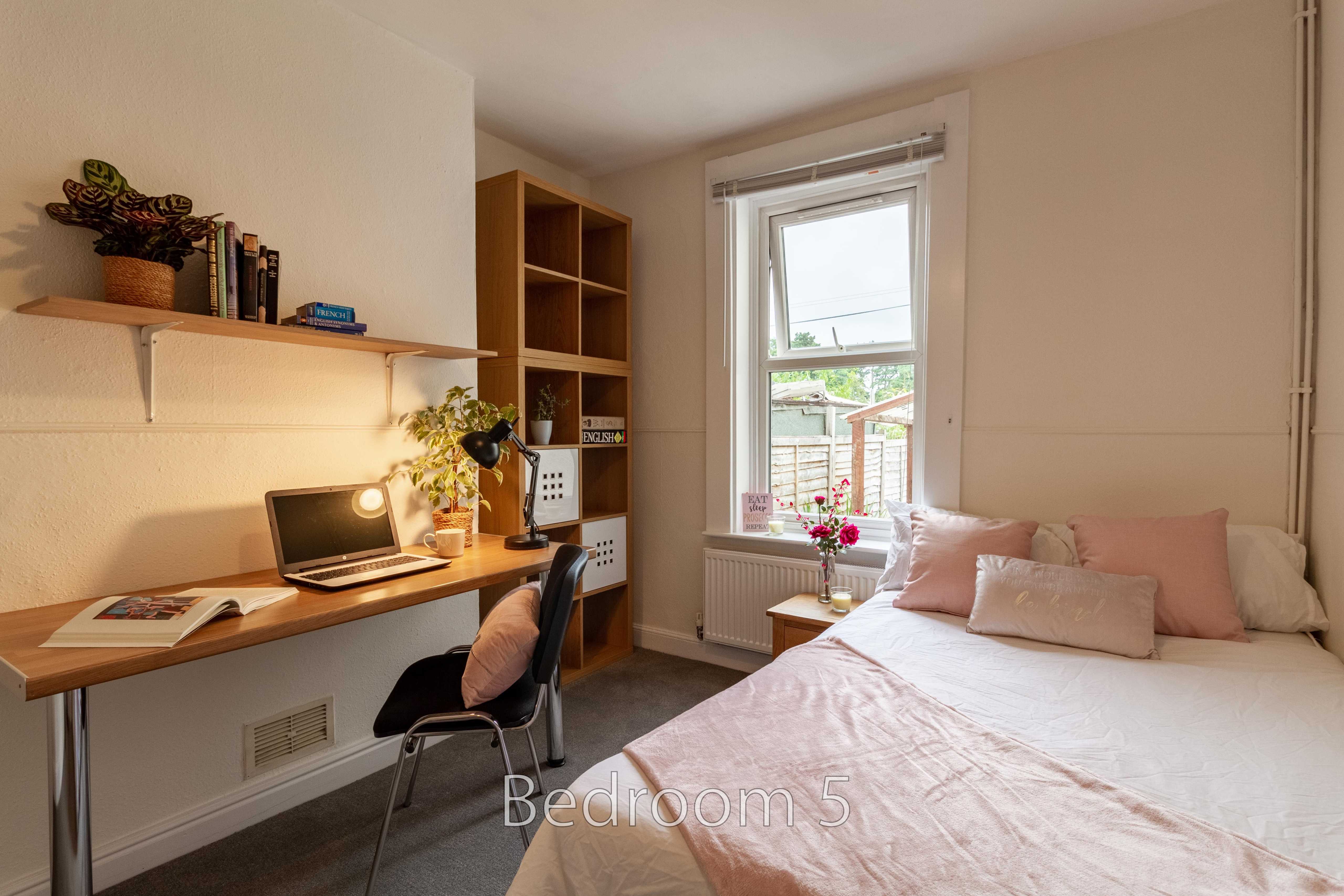 A bedroom with a double bed, desk, chair, shelving and a window.