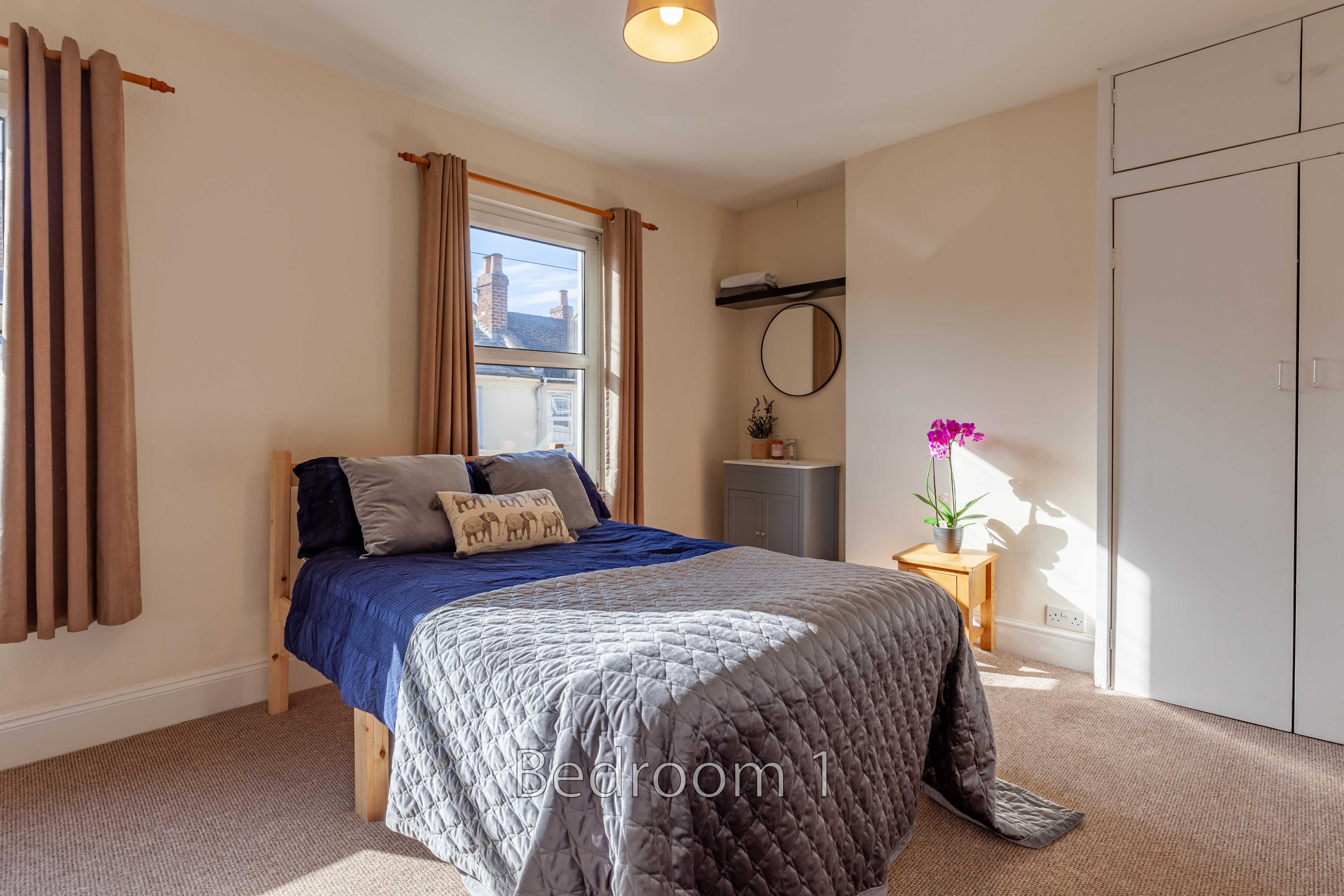 A furnished bedroom with a bed, wardrobe, window, curtains, bedside table and wall mirror.