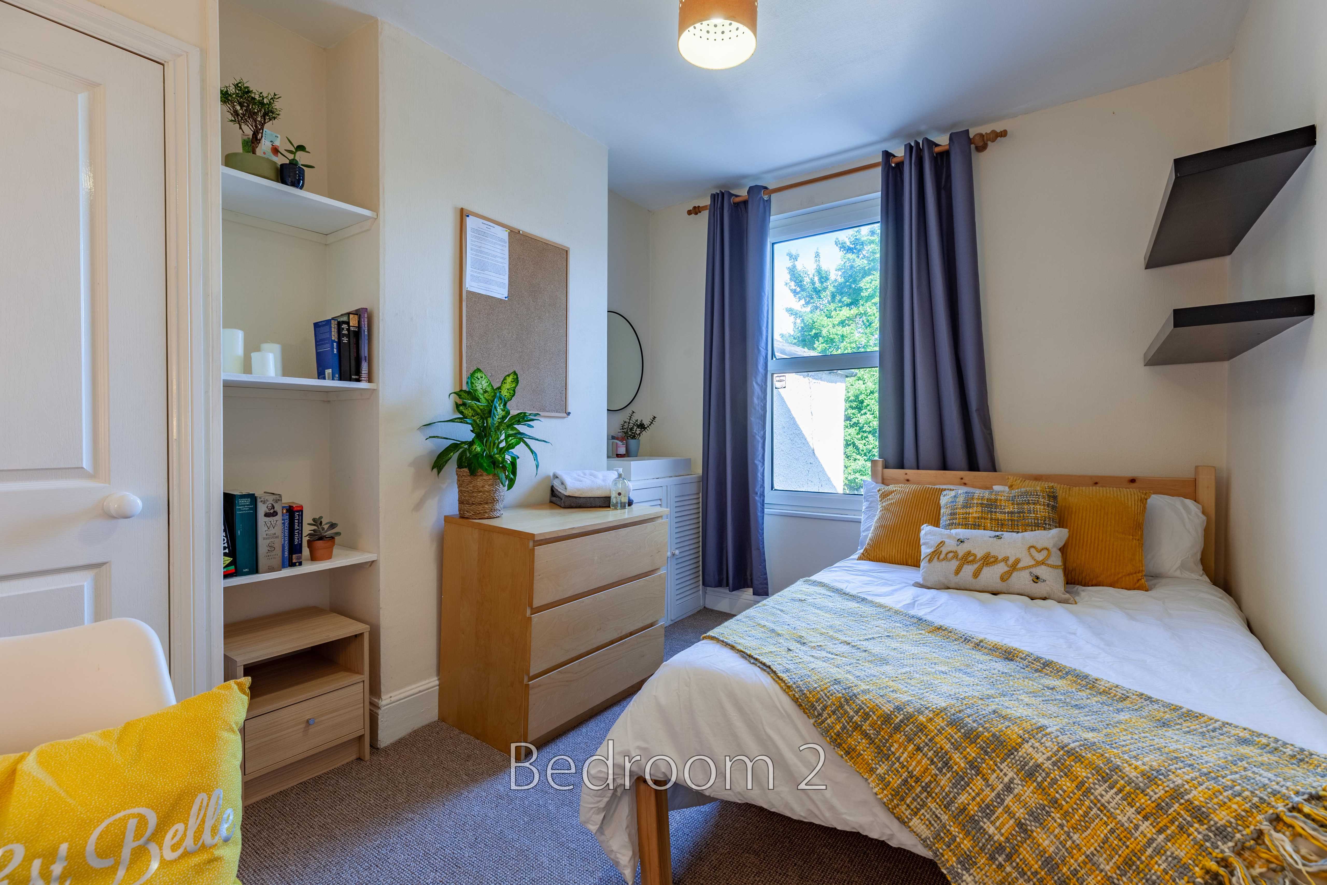 A furnished bedroom with a single bed, window, curtains, shelving and a chest of drawers.