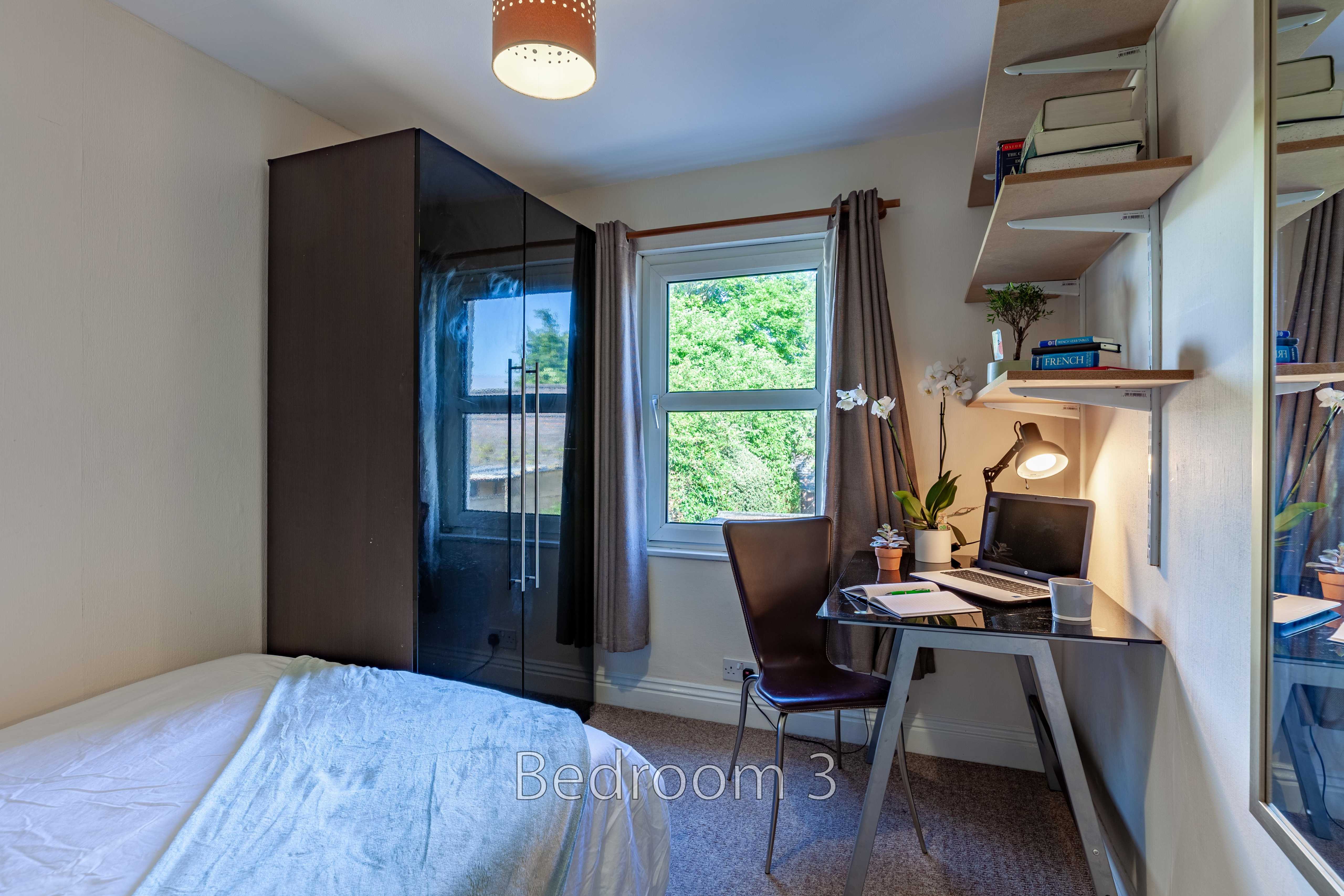 A bedroom with a bed, wardrobe, desk, chair, shelving and a window.