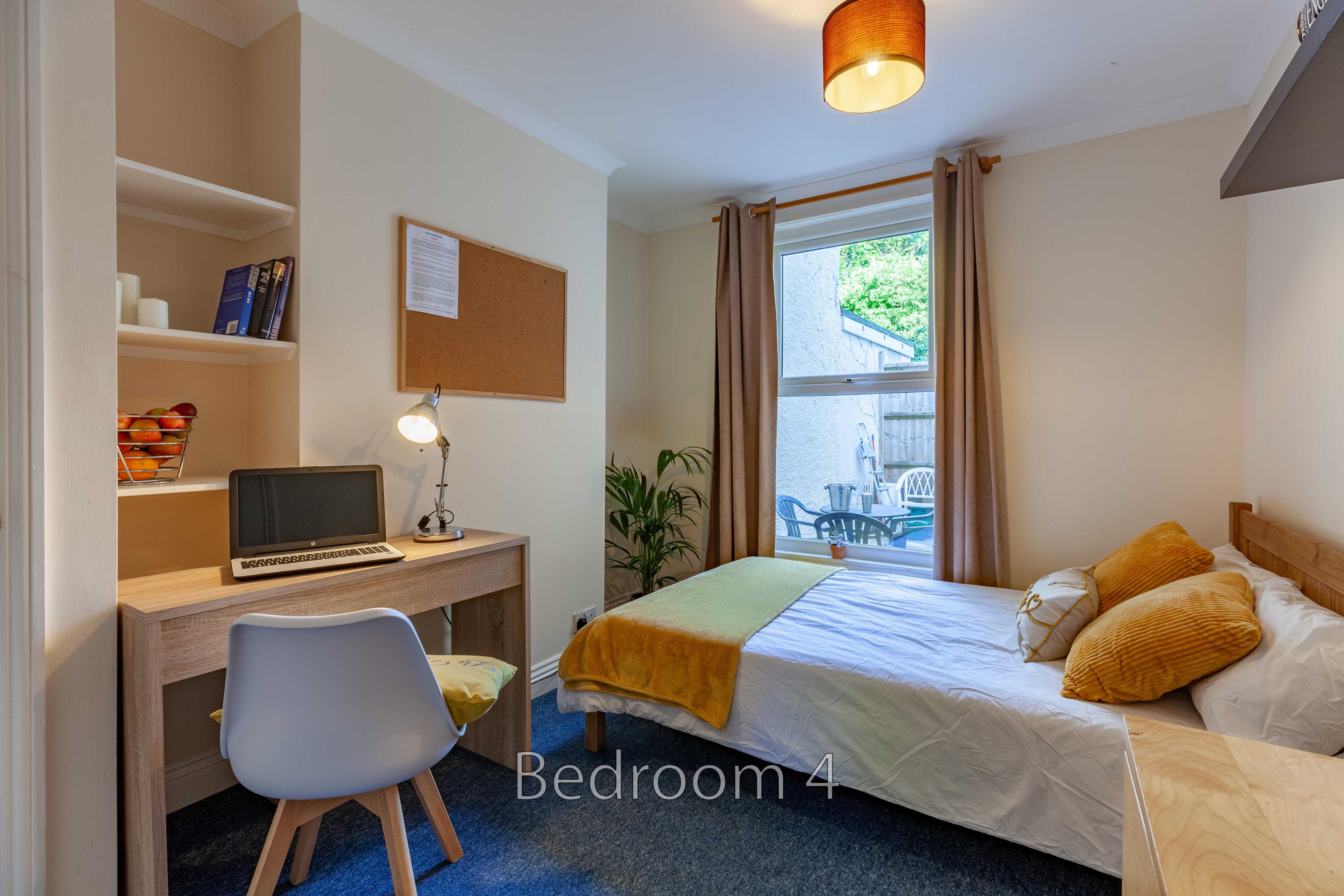 A furnished bedroom with a single bed, desk, chair, shelving, window and curtains.
