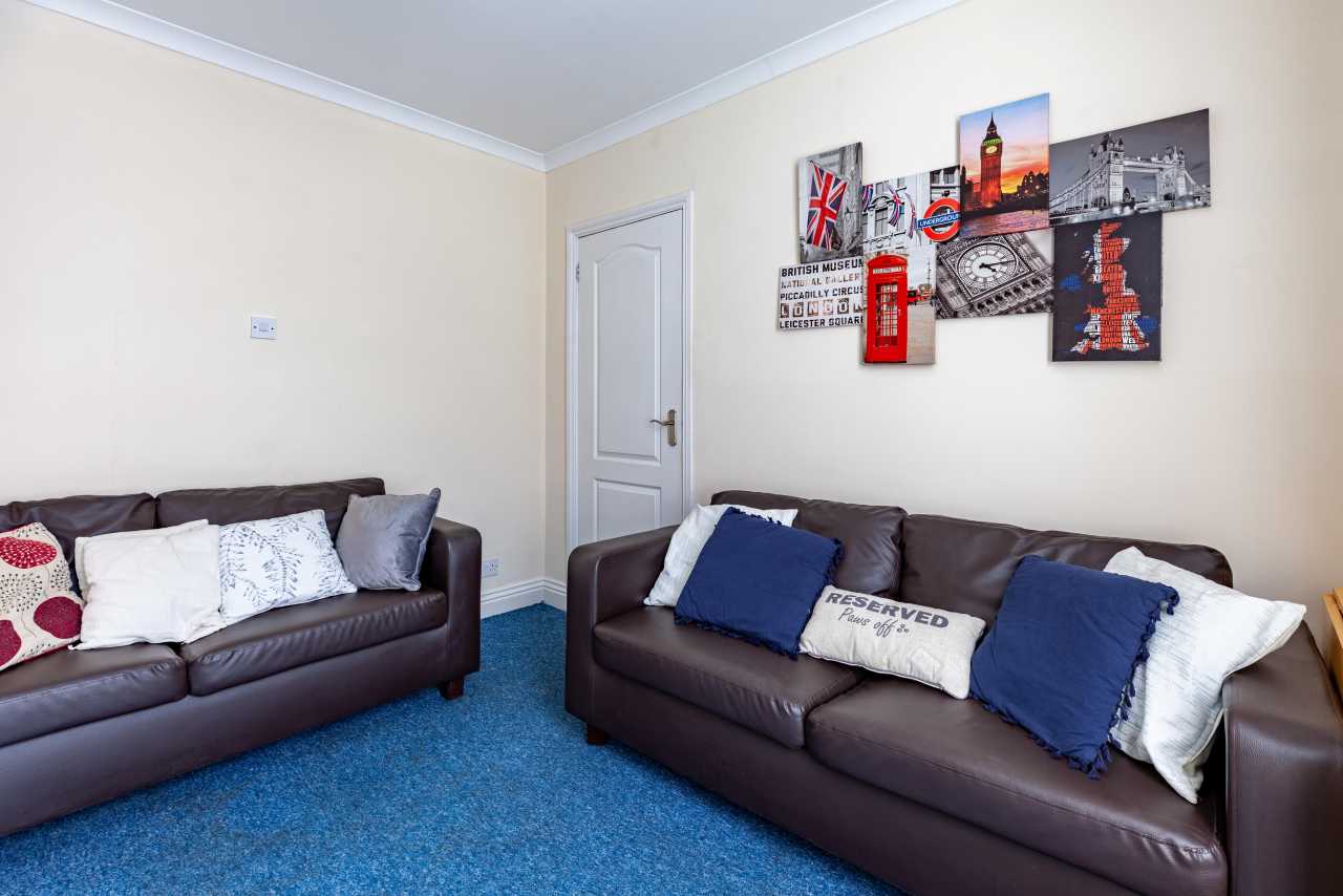 A furnished sitting room with two dark leather sofas, cushions, blue carpet, a closed door and wall art.
