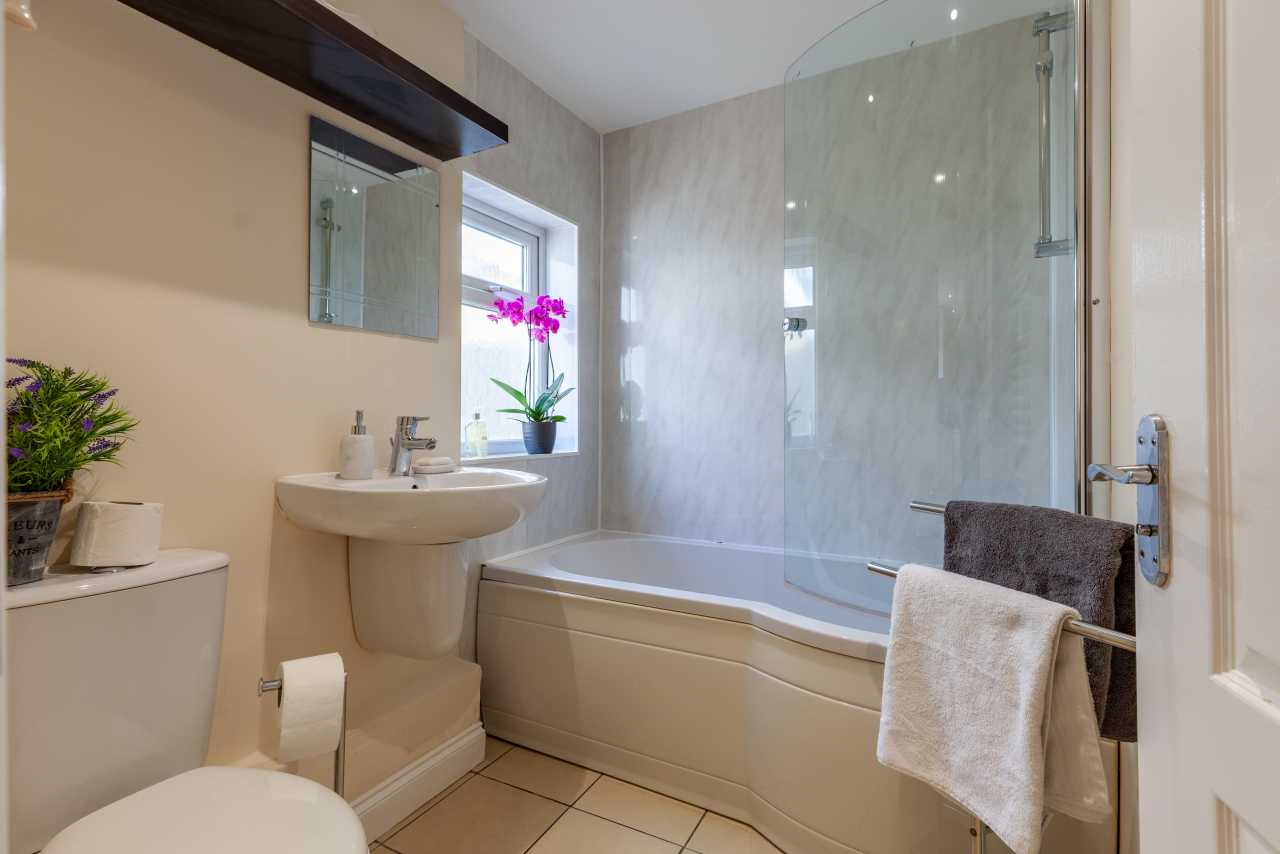 A bright bathroom with a bathtub and glass shower screen, pedestal sink, toilet, tiled floor, mirror, and potted orchid.