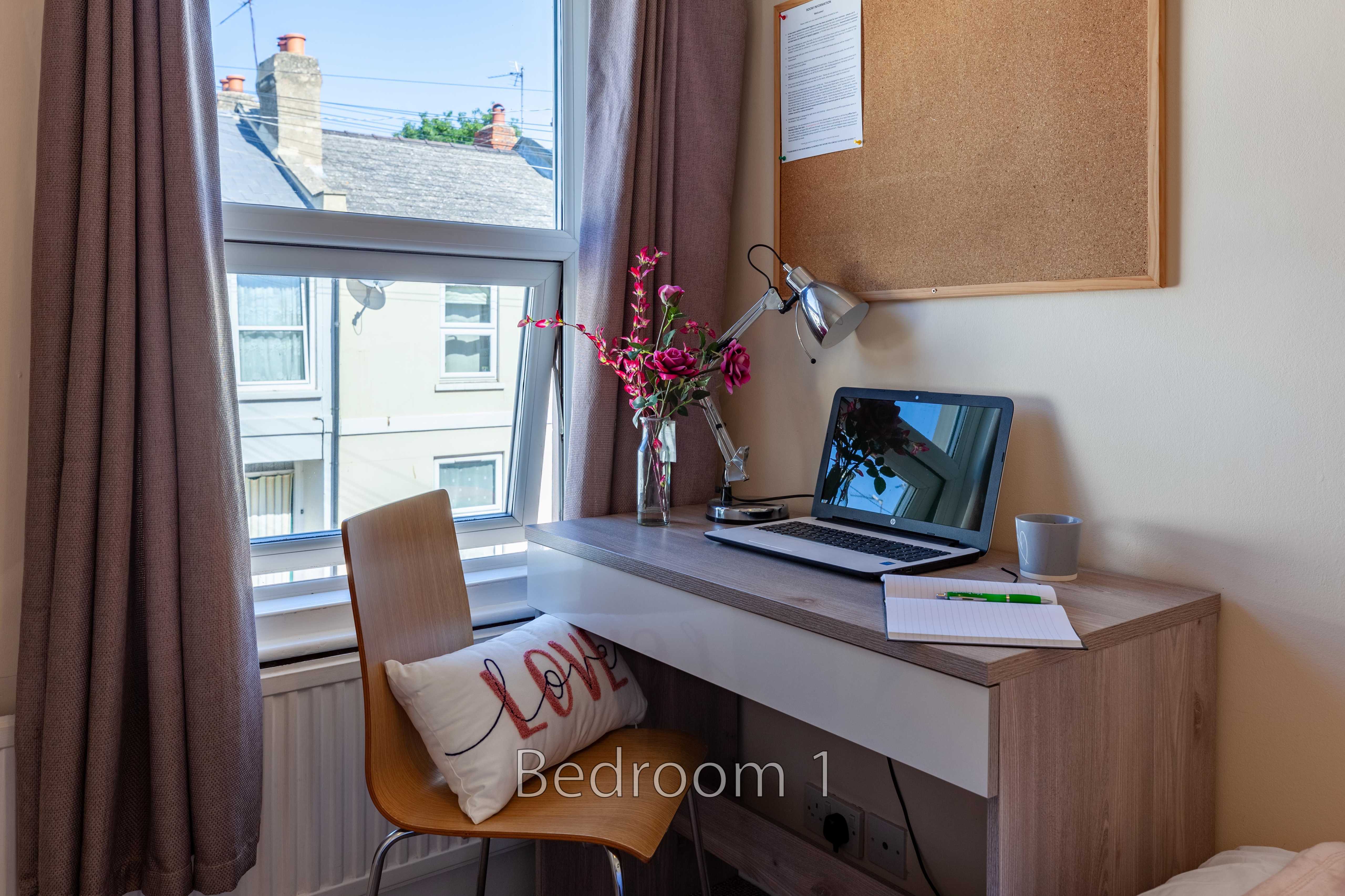 A bedroom with a desk, chair, lamp, laptop, window and bed edge visible.