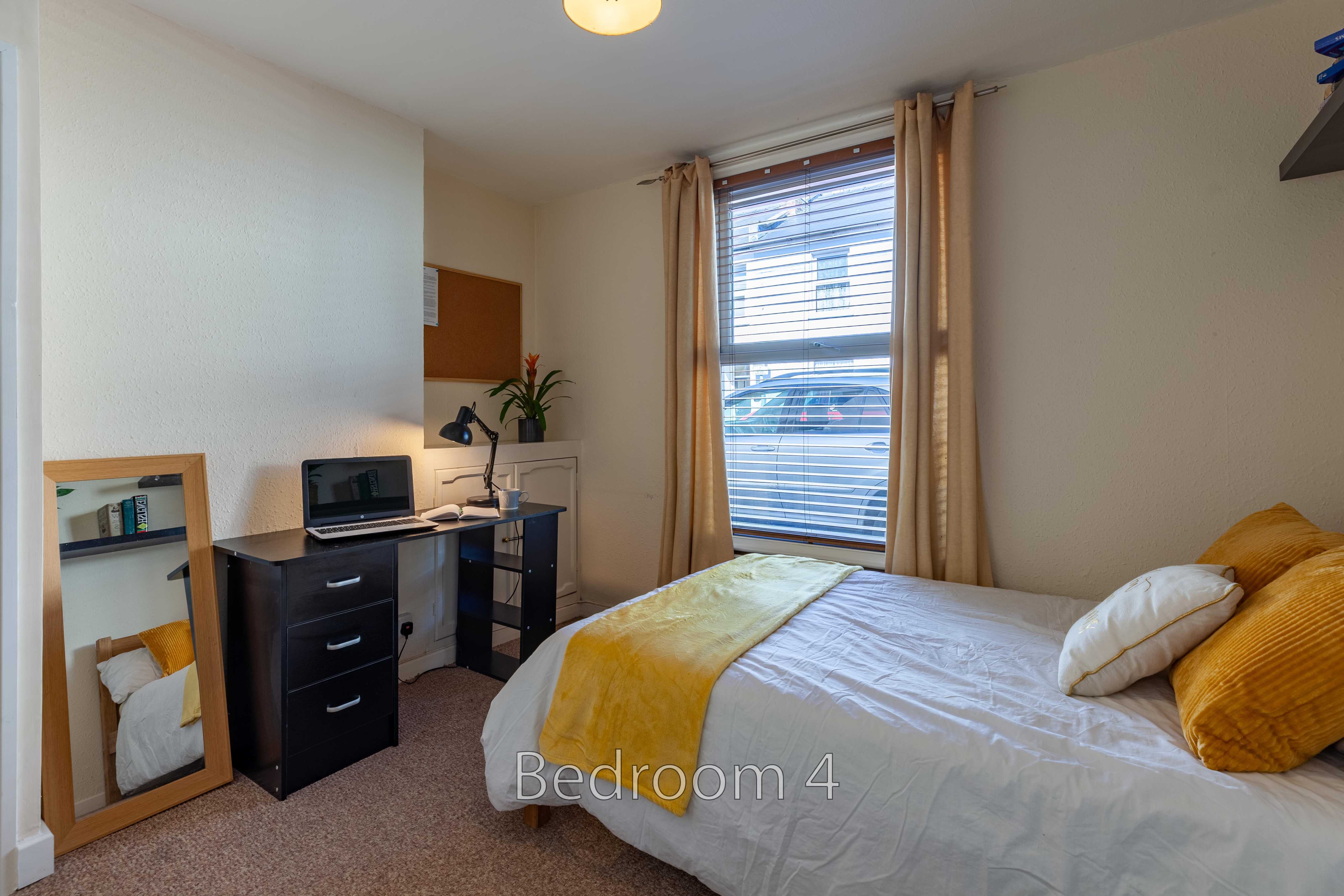 A furnished bedroom with a bed, desk, mirror, window and curtains.