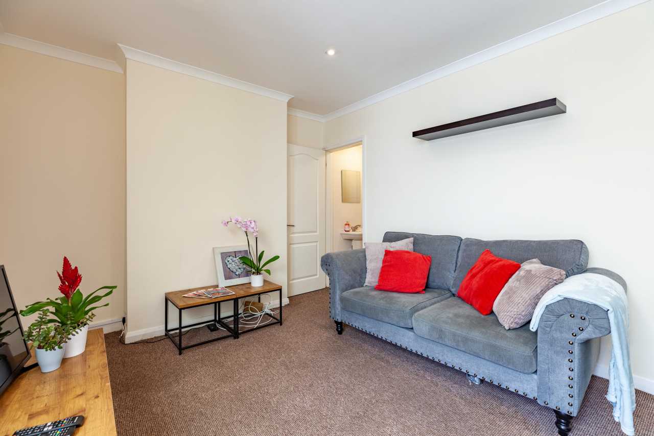 Furnished sitting room with a grey sofa, red cushions, plants, a small coffee table and wall shelf.