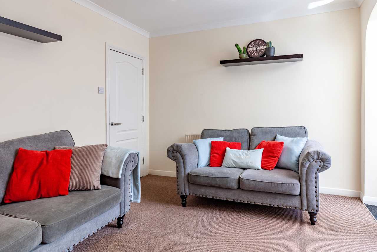 A tidy sitting room with two grey sofas, red and patterned cushions, wall shelves, a closed door and beige carpet.