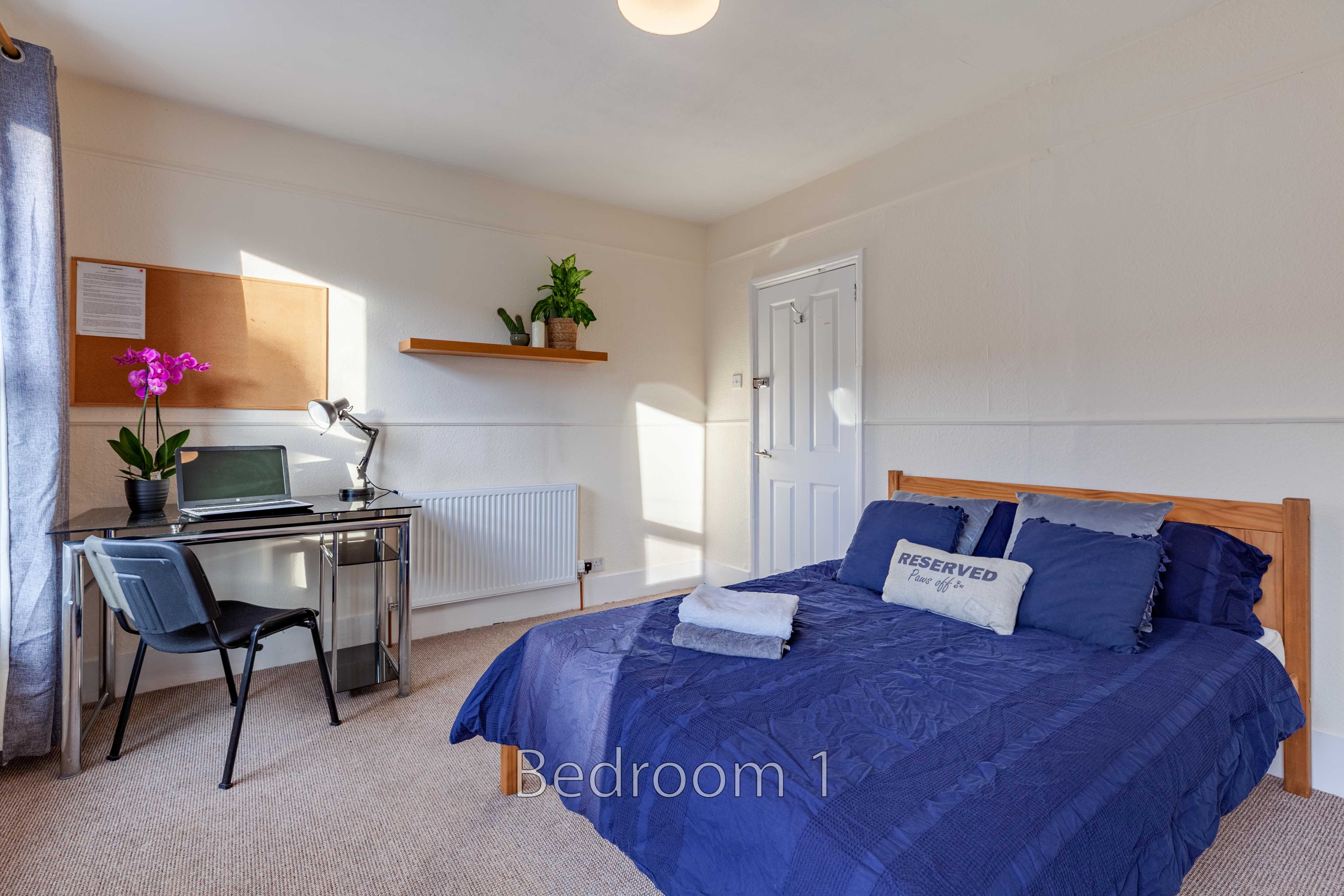 A furnished bedroom with a double bed, desk and chair, radiator, window, and wall shelf.