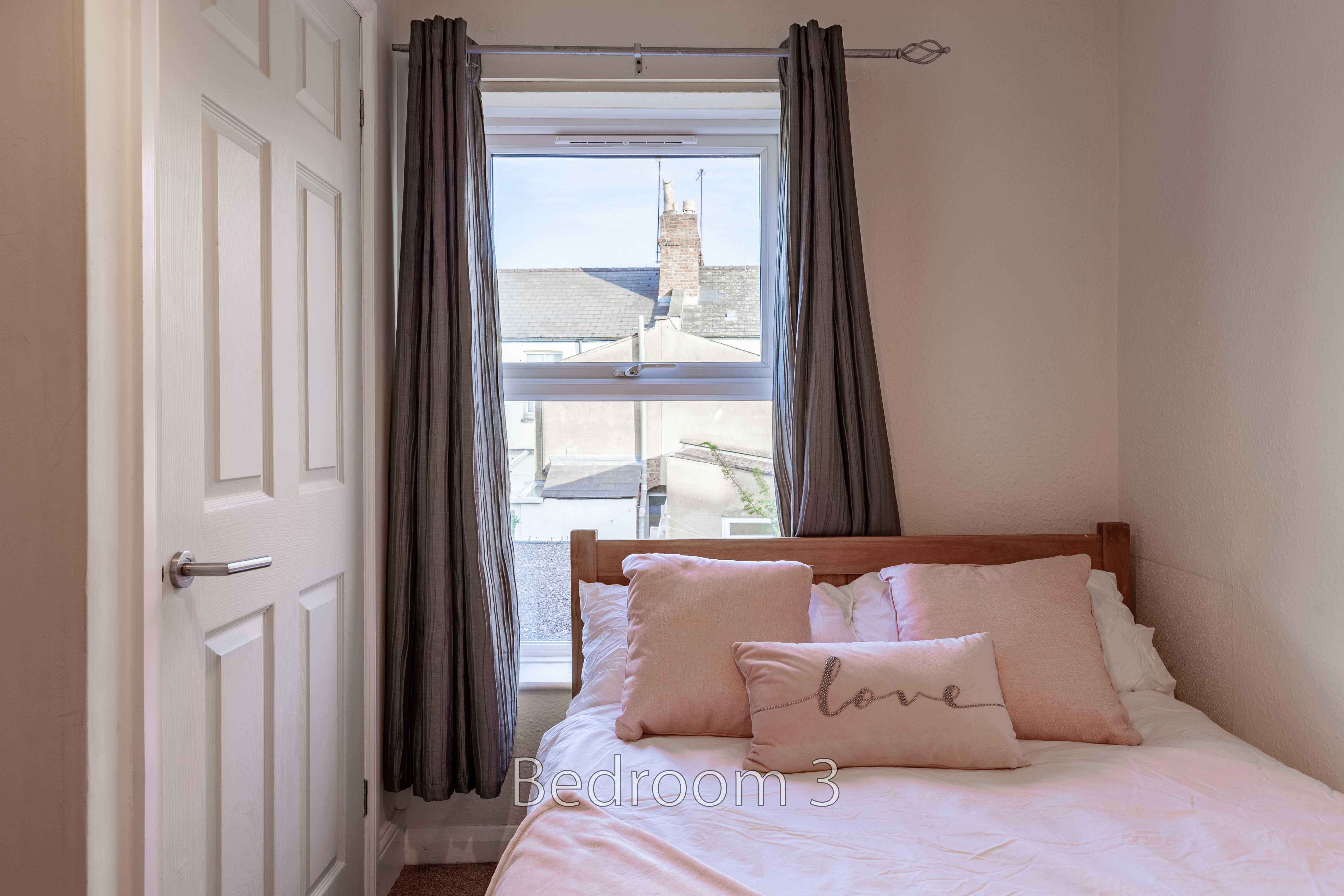 A bedroom with a double bed, pink bedding, curtains, and a window overlooking neighbouring rooftops.