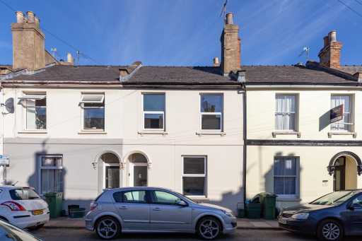 Street view of adjoining two-storey terraced houses with front doors, windows, chimneys, parked cars and wheelie bins.