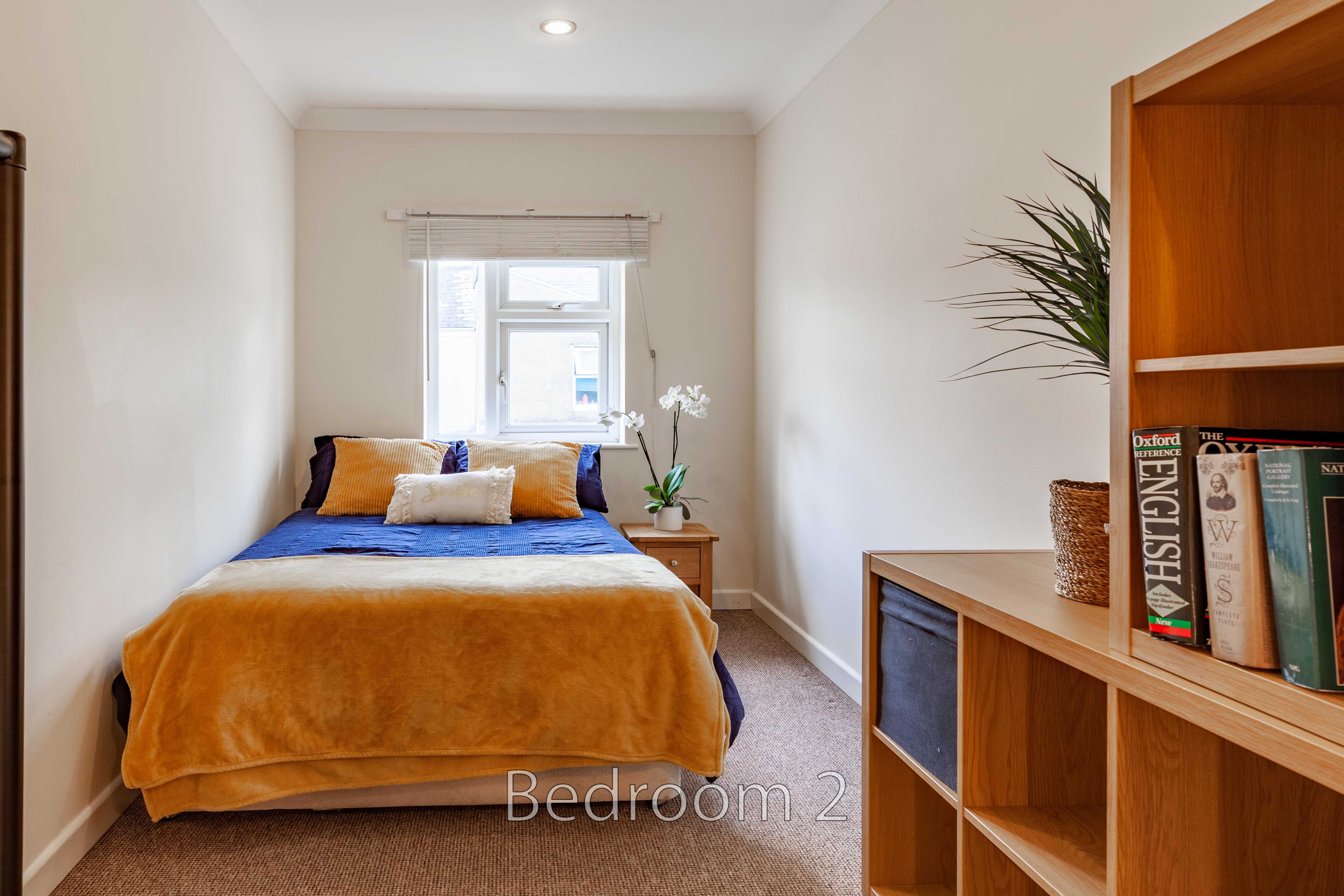 A narrow bedroom with a double bed, blue and mustard bedding, a window, carpeted floor and wooden shelving.