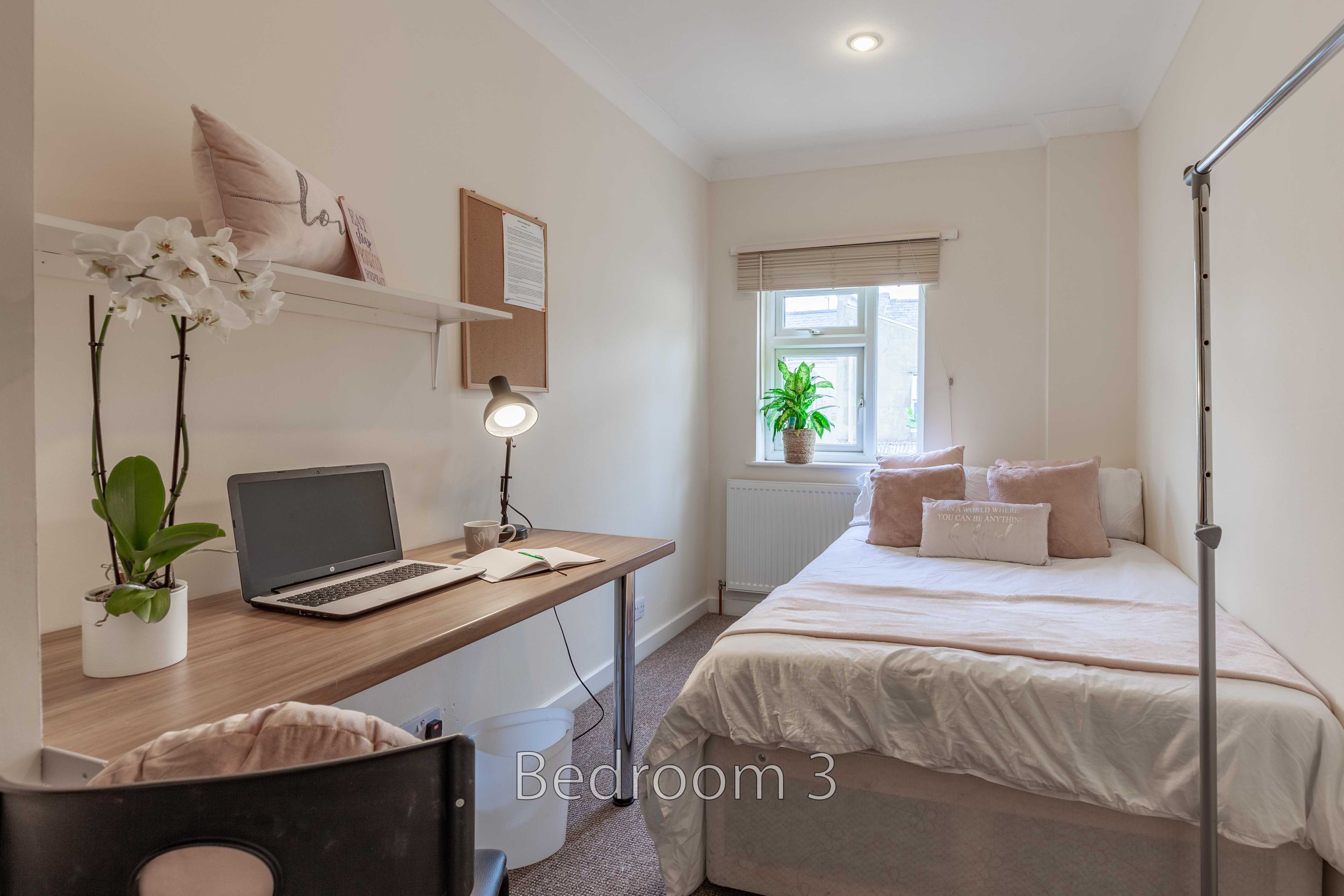 A furnished bedroom with a double bed, desk, laptop, chair, shelves, and a window.