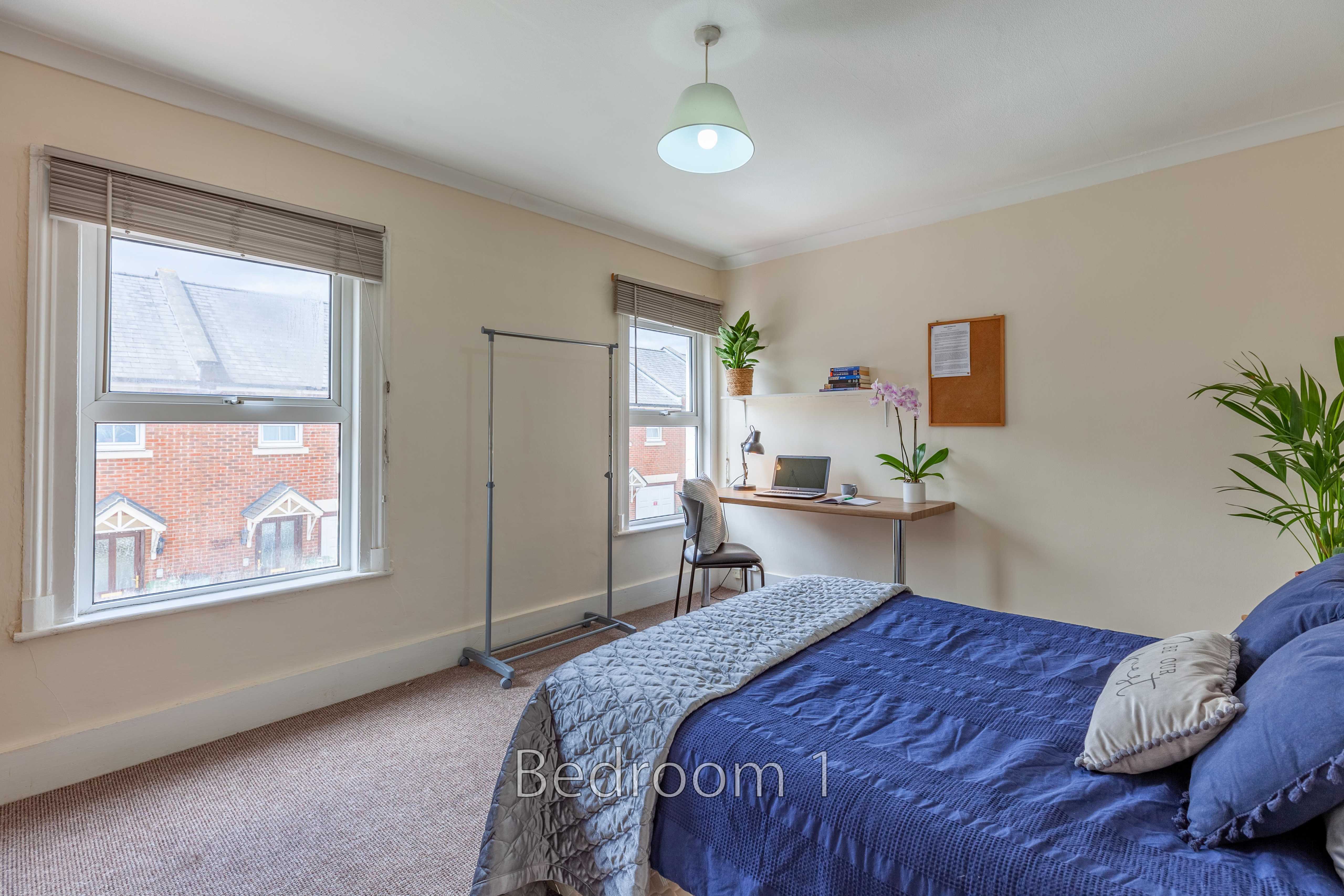 A furnished bedroom with a double bed, desk, chair, windows, and potted plants.