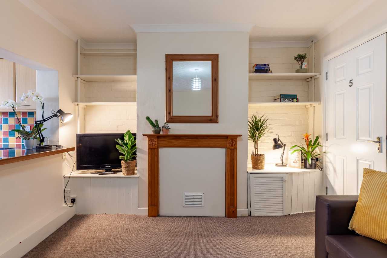 Bright furnished communal sitting room with a fireplace, built-in shelving, television, plants and a door labelled 5.