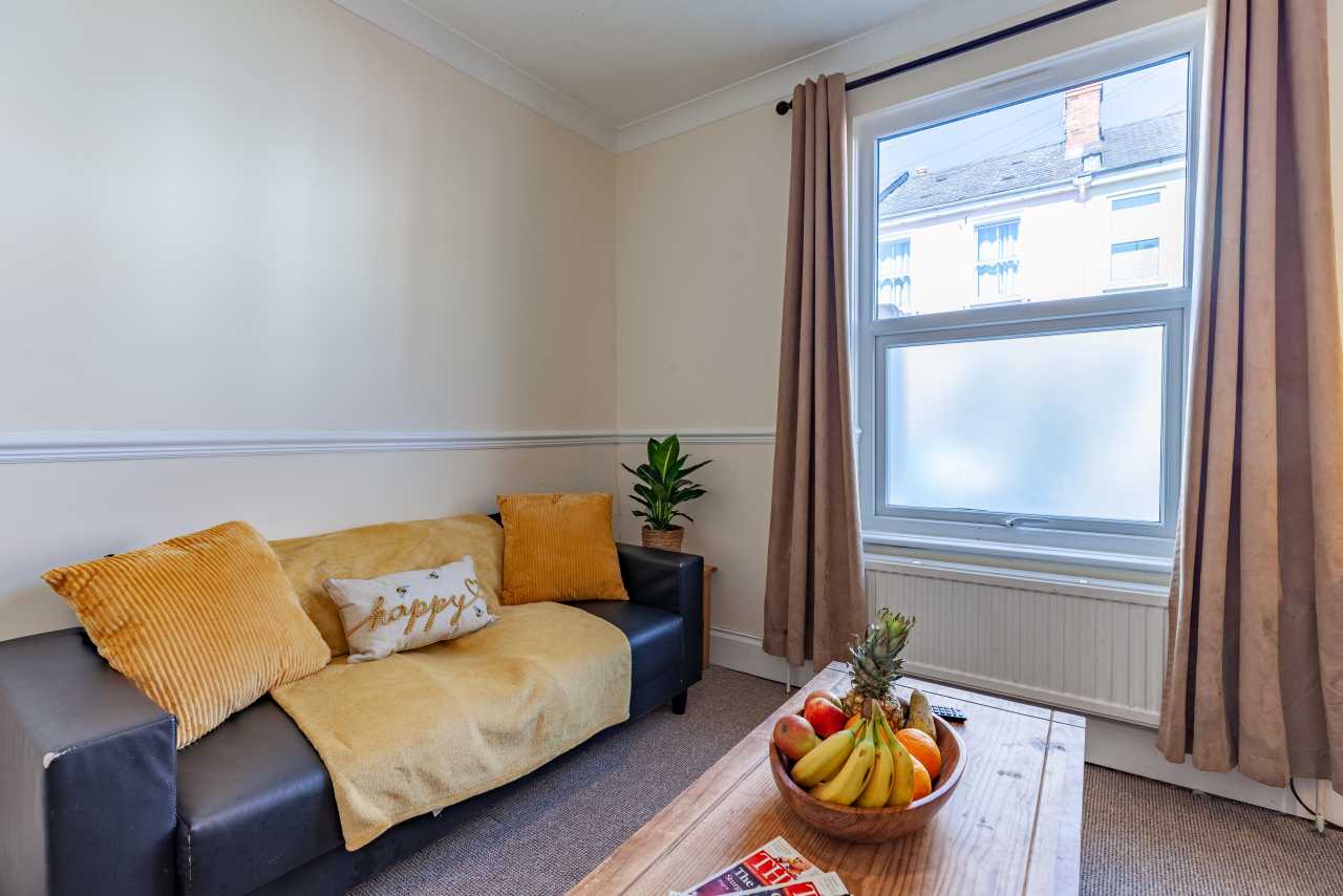 A furnished sitting room with a sofa, coffee table, fruit bowl, curtains and a window.