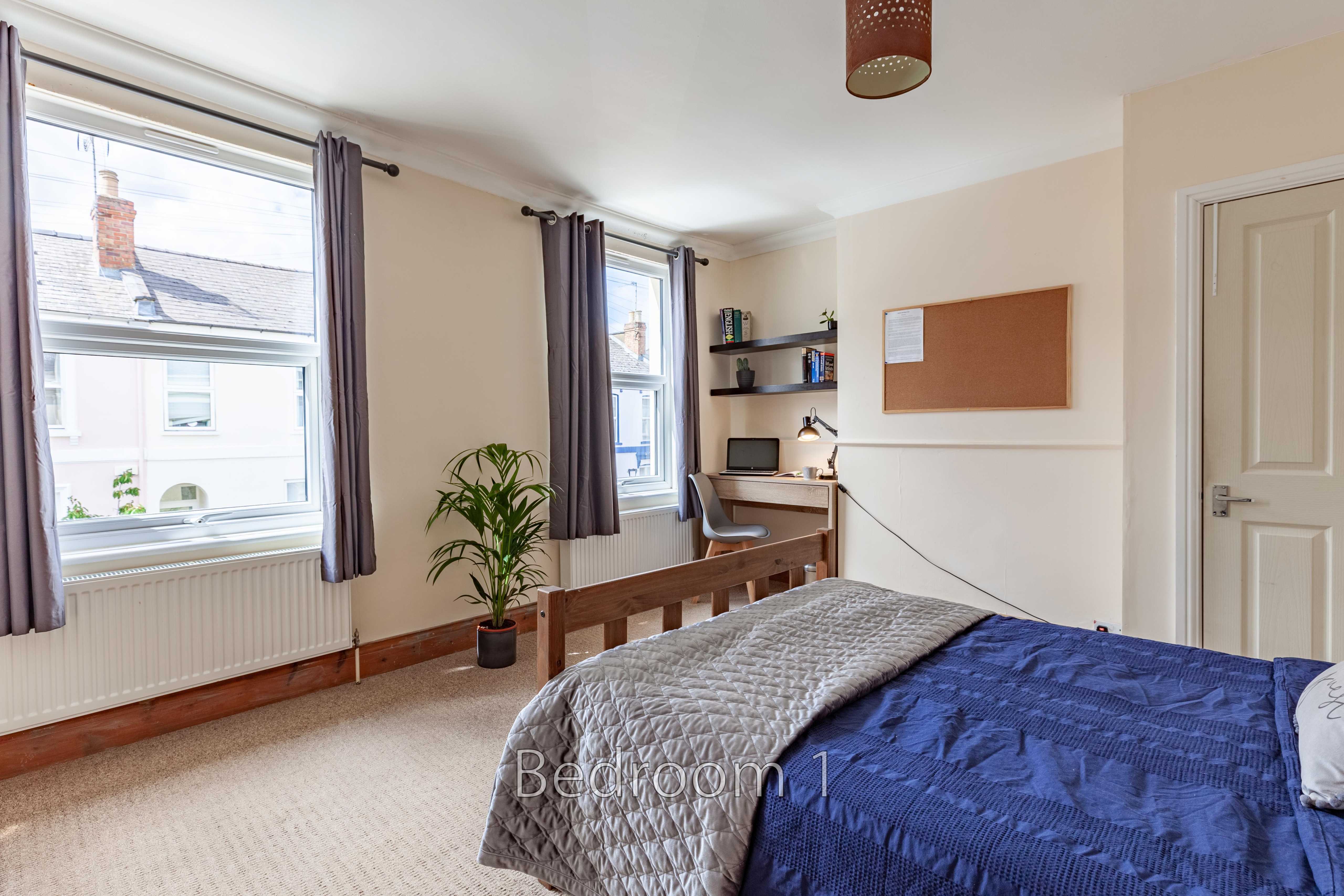 A bedroom with a bed, two windows, a desk, shelving and a potted plant.