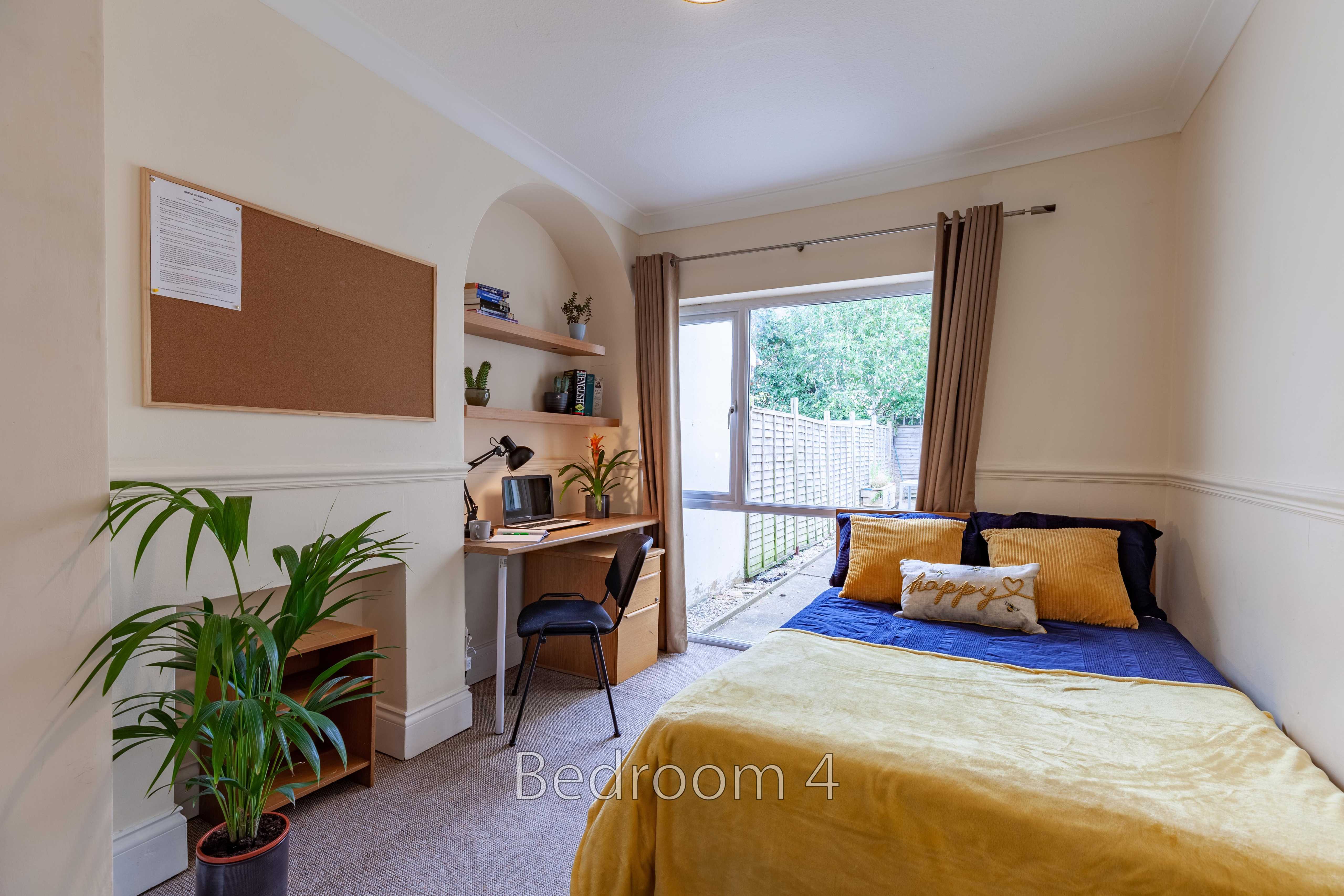 A furnished bedroom with a double bed, desk, shelving, plants and glass doors opening to an outdoor area.