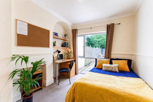 A furnished bedroom with a double bed, desk, shelving, plants and glass doors opening to an outdoor area.