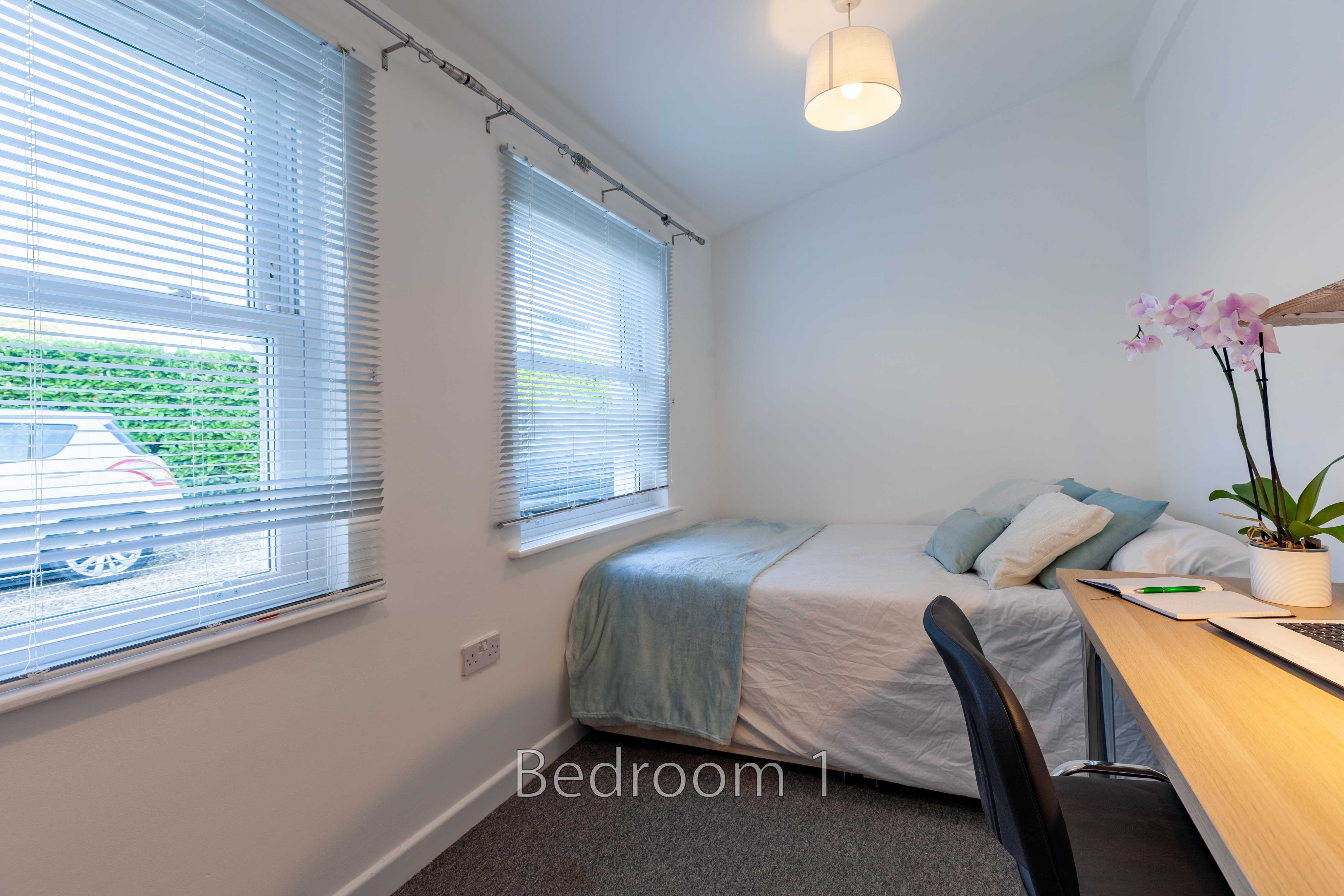 A furnished bedroom with a single bed, desk, chair, windows with blinds, and a potted orchid.