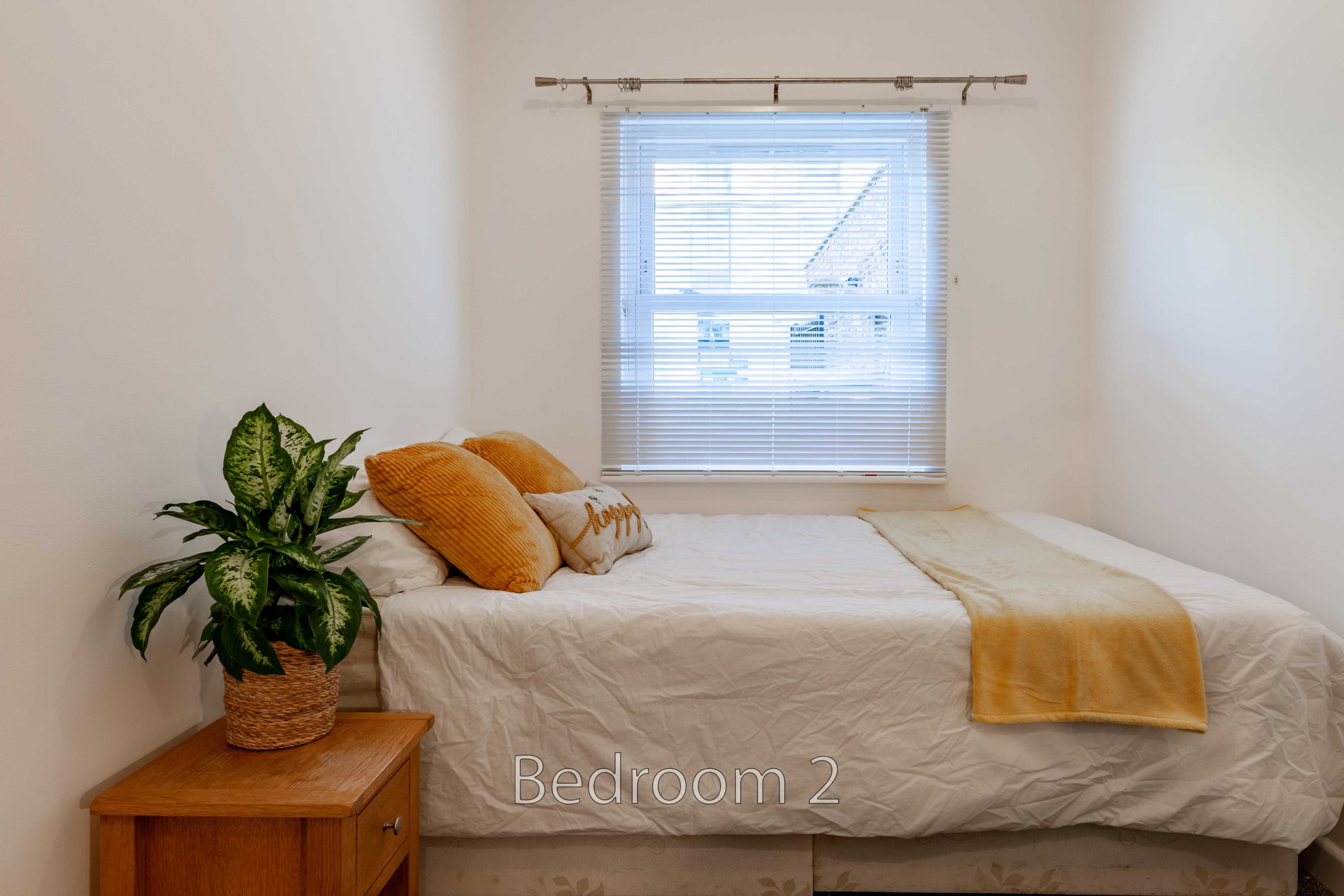 A bedroom with a double bed, orange cushions, a yellow throw, a window with blinds, and a potted plant.