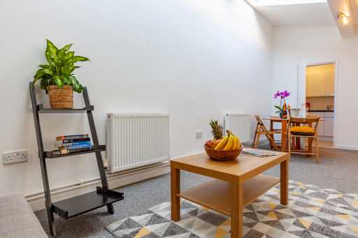 Bright furnished living room with a coffee table, patterned rug, shelving, plants, dining table and an open doorway.