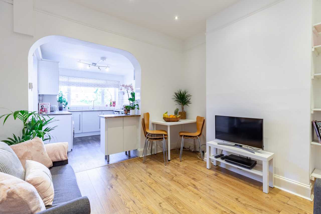 Bright open-plan living and dining area with a television, dining table, kitchen, sofas and potted plants.