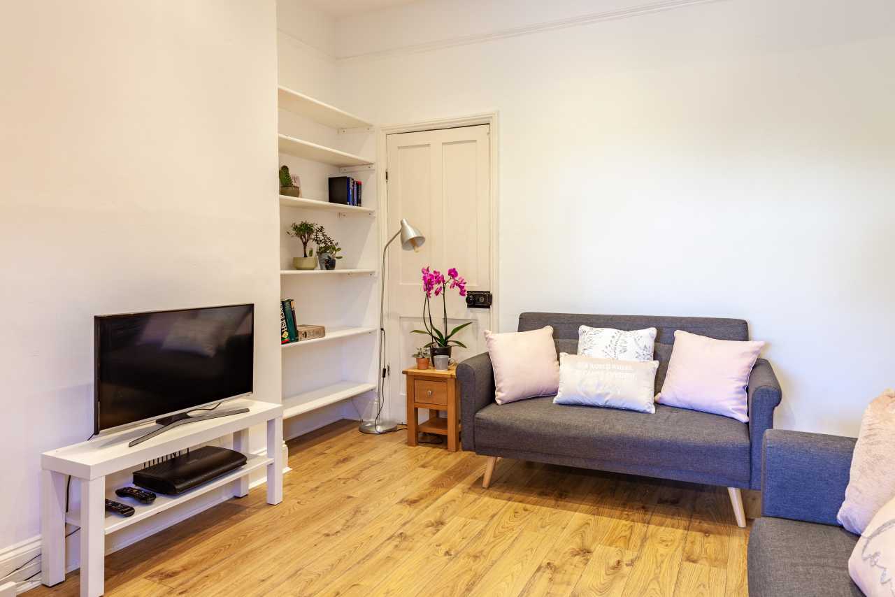 Bright living room with a television, shelving, sofa, armchair, side table and wooden flooring.