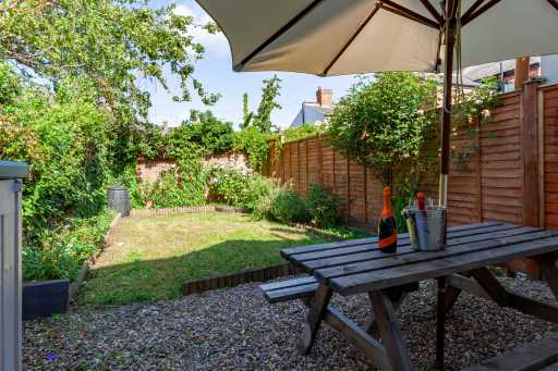 A fenced garden with grass, plants, gravel, and a wooden picnic table beneath a large umbrella.