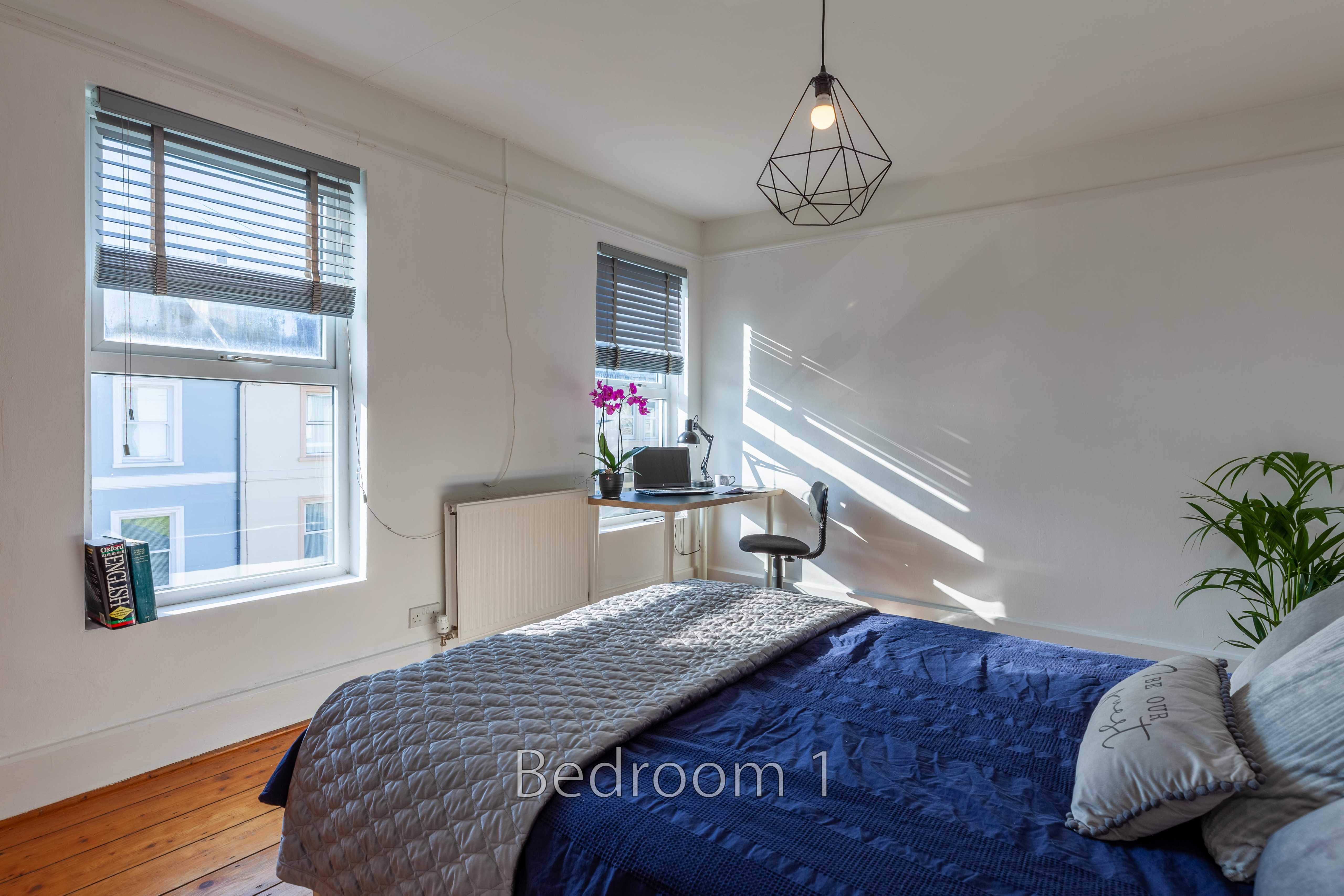 A bedroom with a double bed, two windows, a desk, hanging light and potted plant.