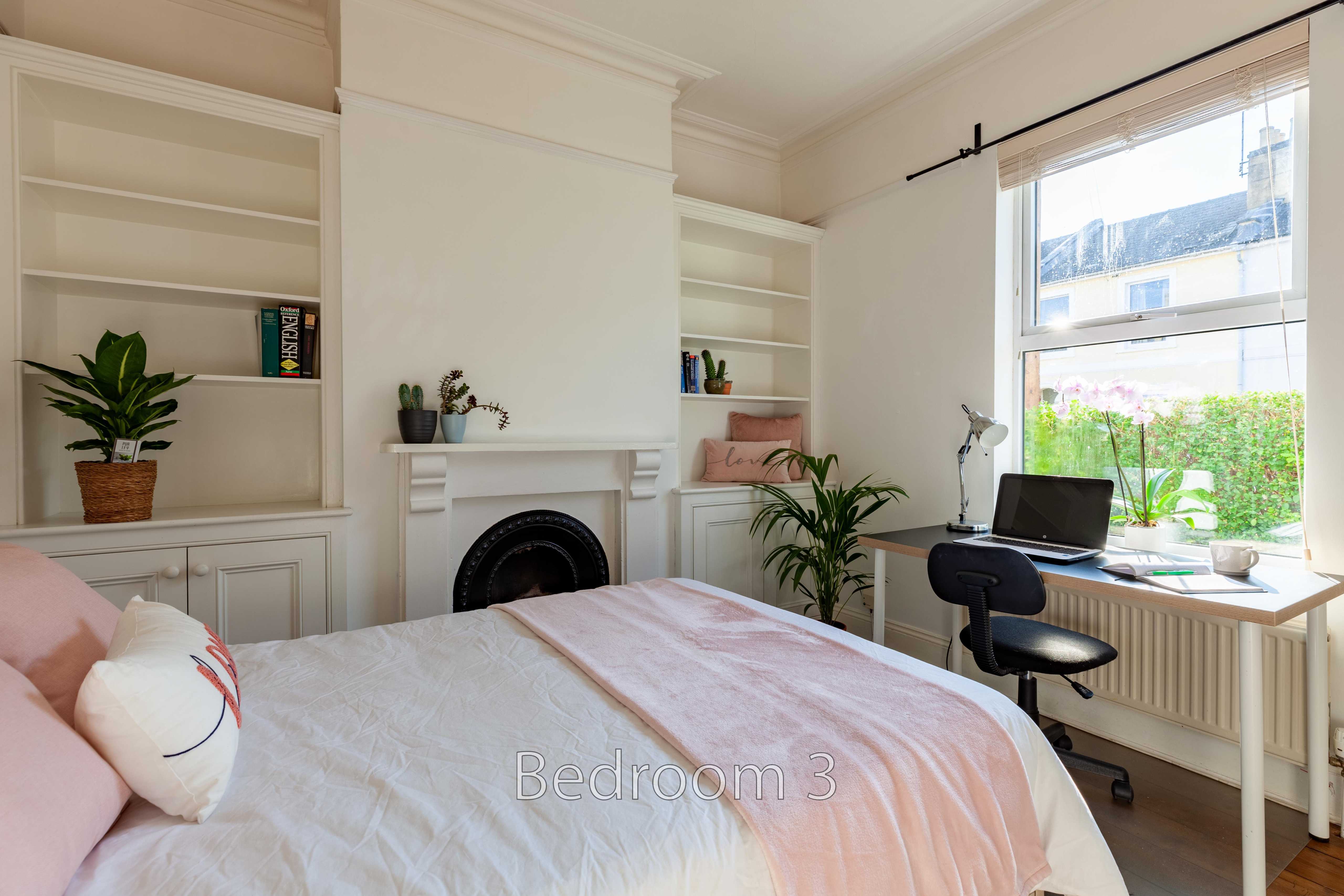 A bright bedroom with a double bed, fireplace, built-in shelving, desk and large window.