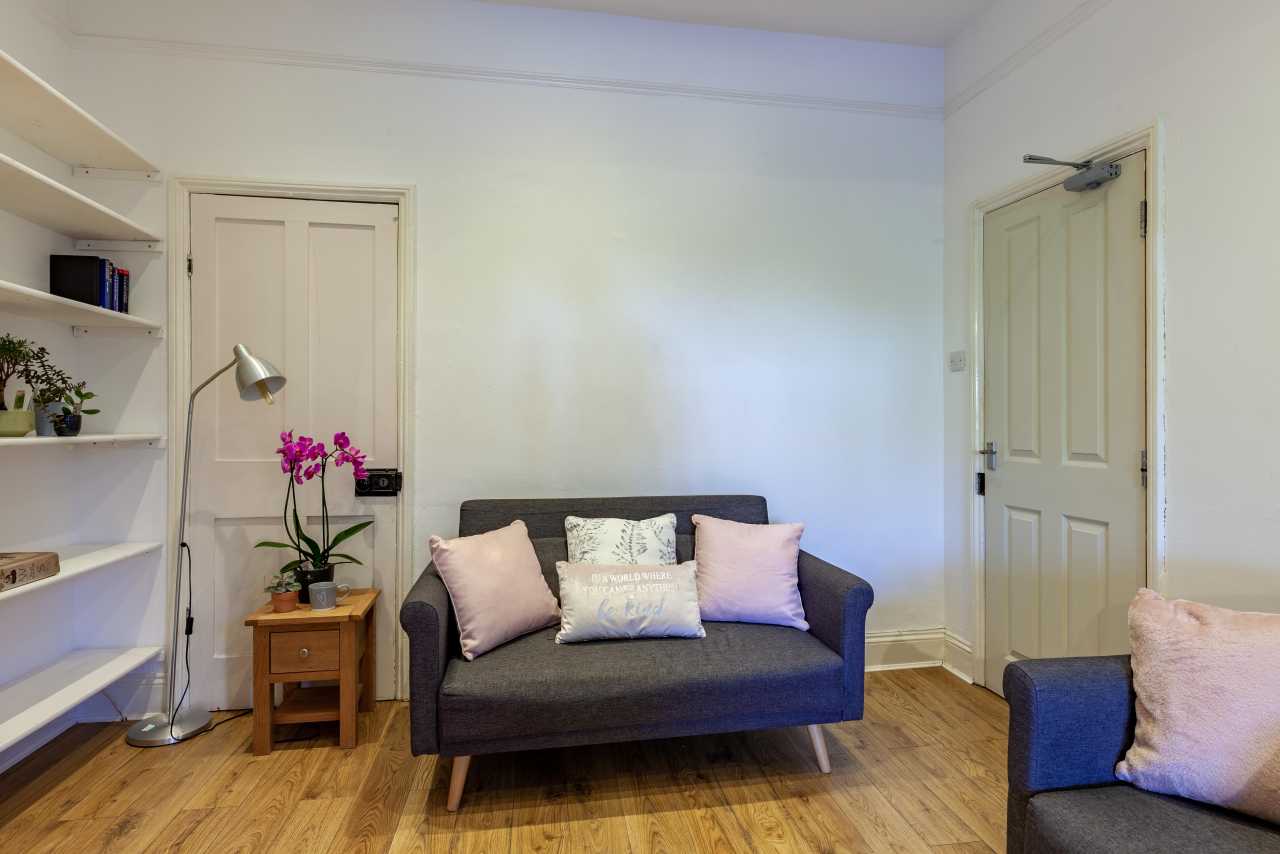 A furnished room with a sofa, armchair, side table, floor lamp, shelving and wooden flooring.